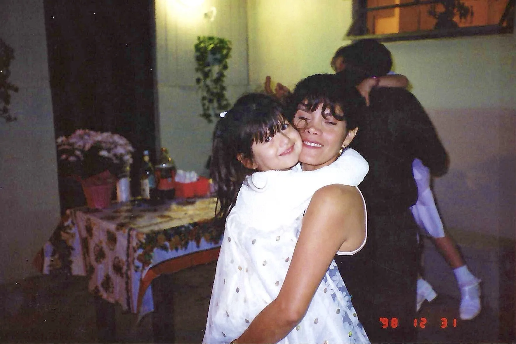 A woman with short, curly dark hair holding a young girl with dark hair, both smiling and hugging each other at a party or gathering.
