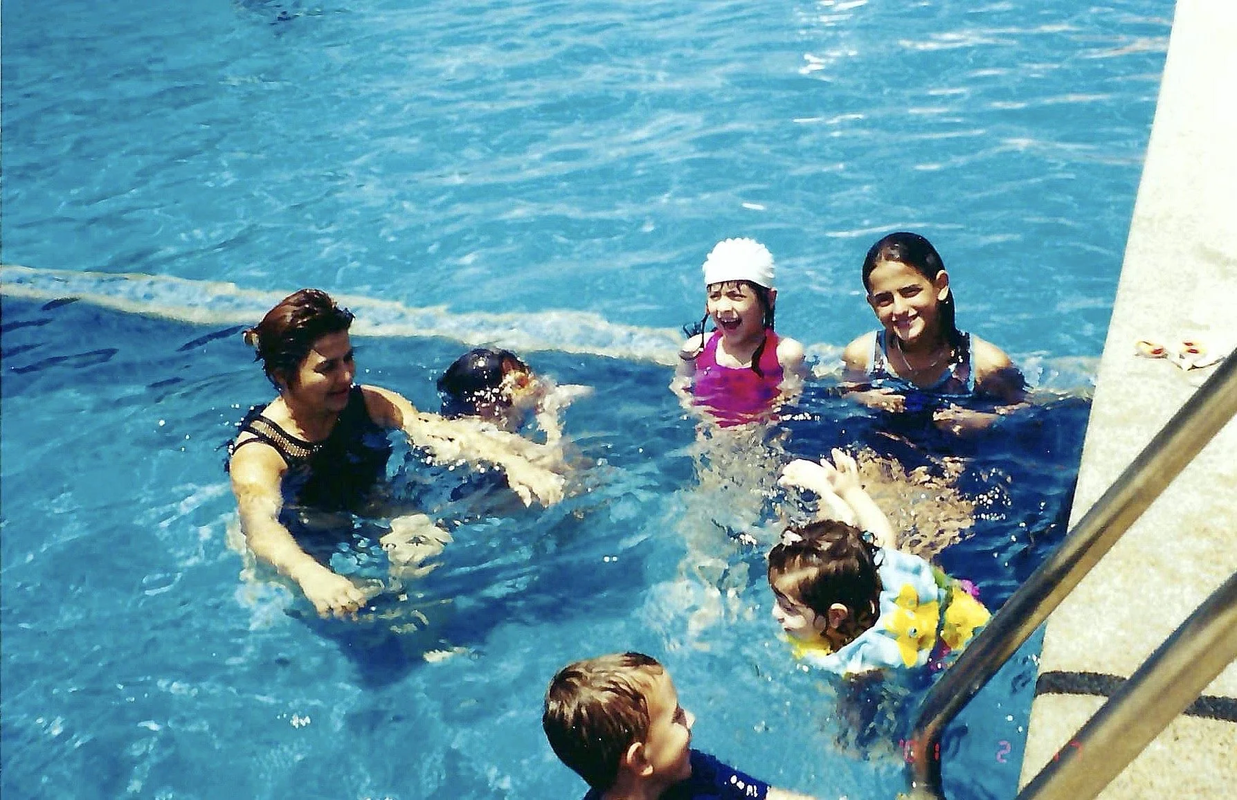 Group of children and adults swimming and playing in a swimming pool, smiling and enjoying the water.