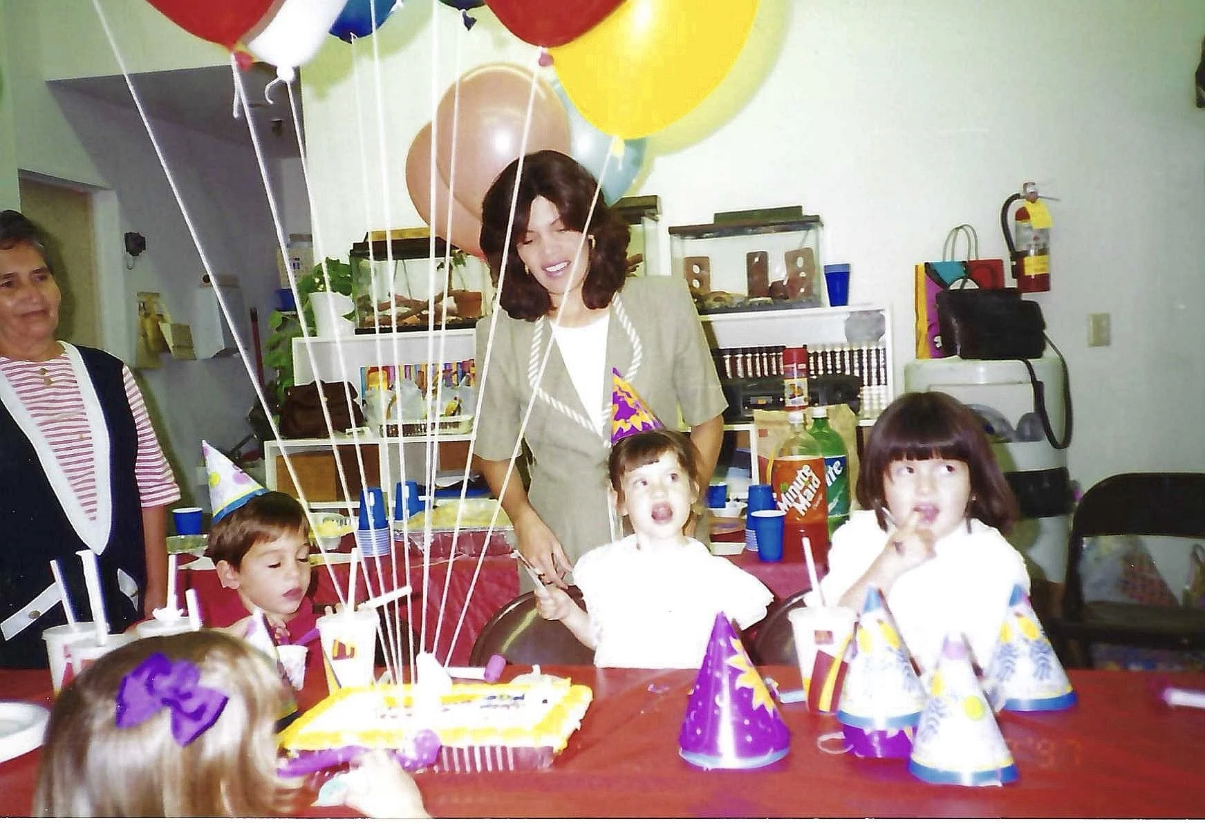 Children celebrating birthday party with balloons, hats, cake, and party favors at a decorated indoor venue.