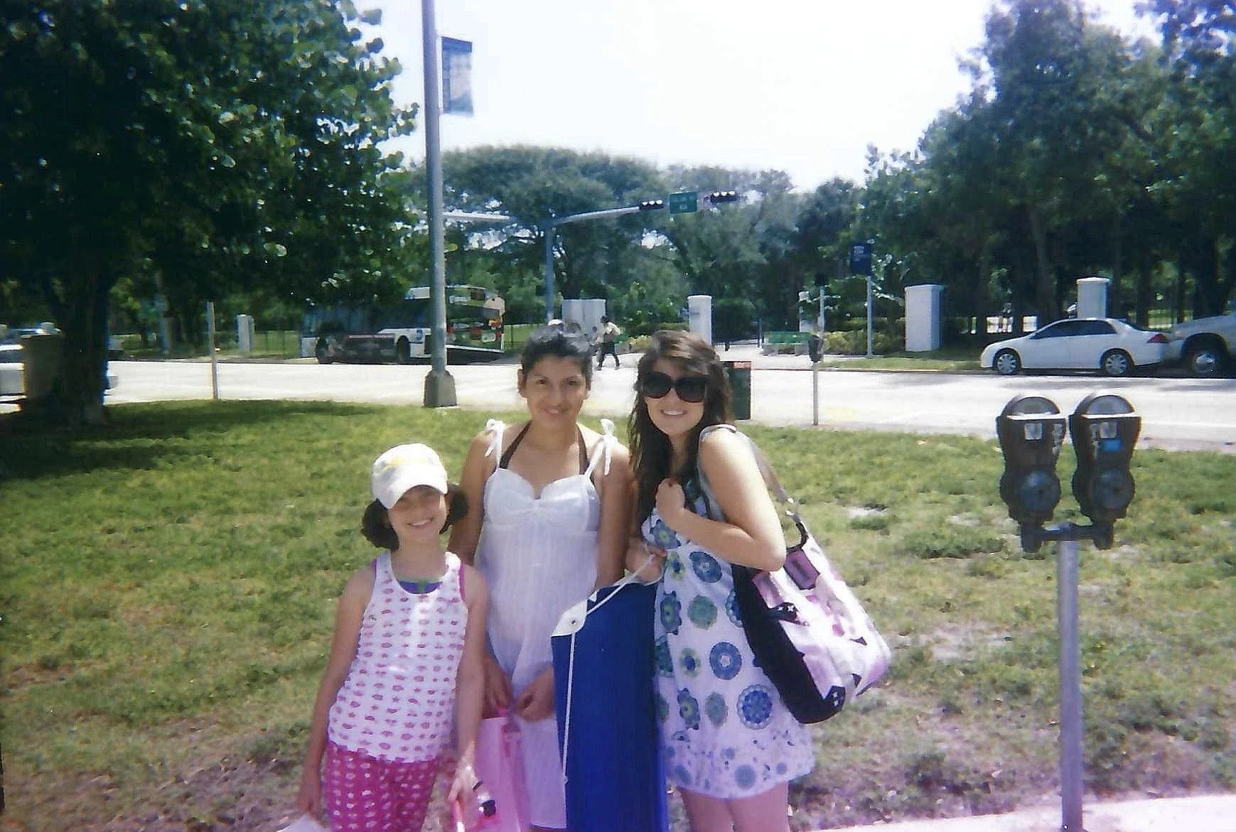 Three females, two women and one girl, standing outdoors in front of a park, smiling at the camera. The girl is wearing a white hat and a pink dress, one woman is wearing sunglasses and a patterned dress, and the other woman is in a white sundress wi