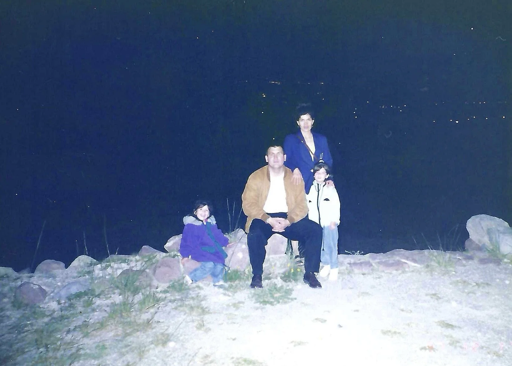 A family of four posing outdoors at night on rocky terrain with a starry sky in the background. The man is seated, wearing a brown jacket, and the woman and children are standing beside him.