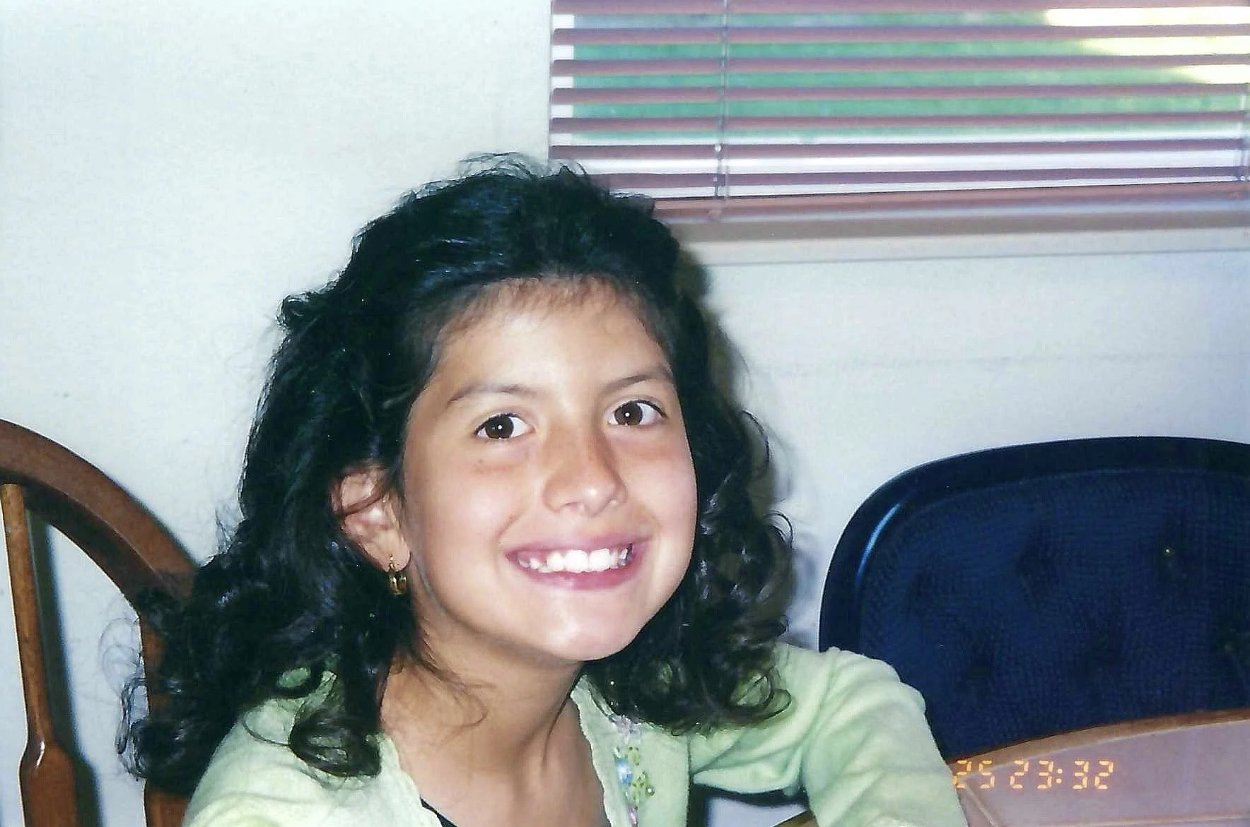 A young girl with dark, curly hair and a bright smile, wearing a light green top, sitting indoors near a window with closed blinds.