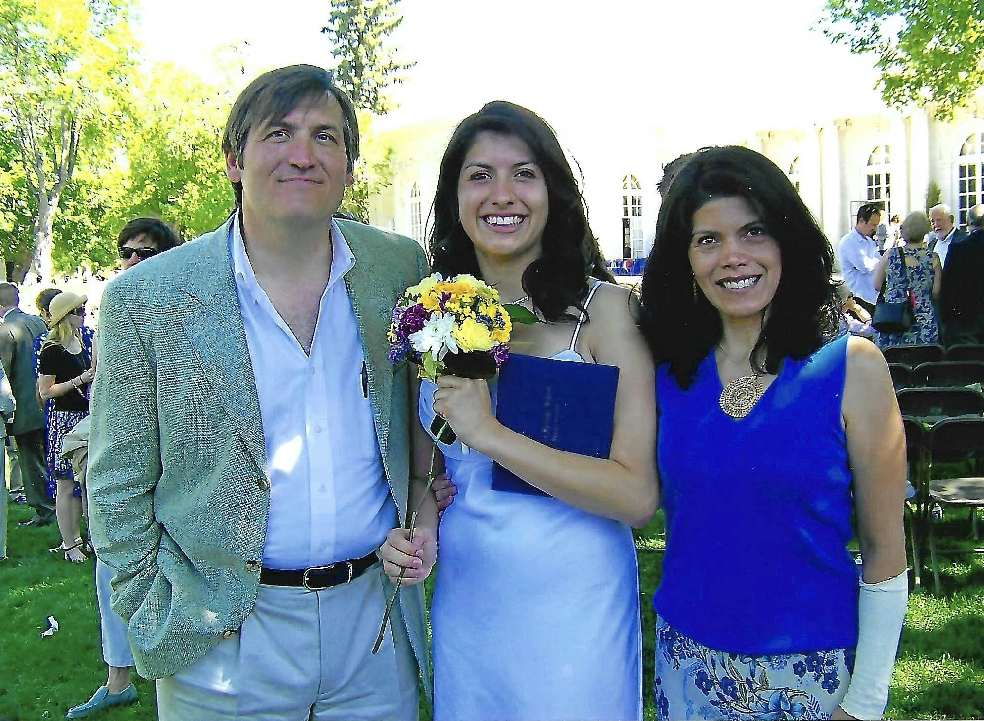 Three people are standing outdoors during a sunny day, with green trees in the background. The person on the left is a man wearing a shiny light blue blazer and a white shirt. The person in the middle is a woman holding a bouquet of colorful flowers 