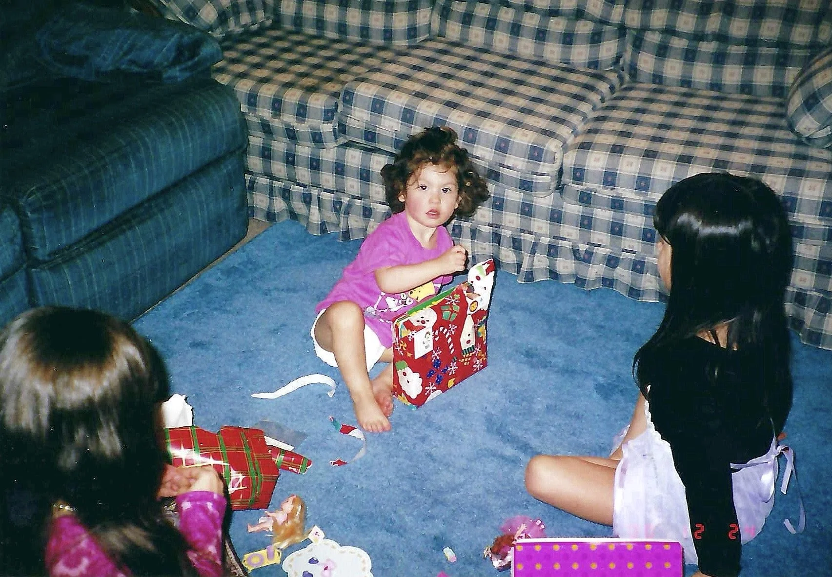 A young girl with curly hair in a pink shirt and white shorts sitting on a blue carpet, opening Christmas presents with other children around her, in a living room with plaid couches.