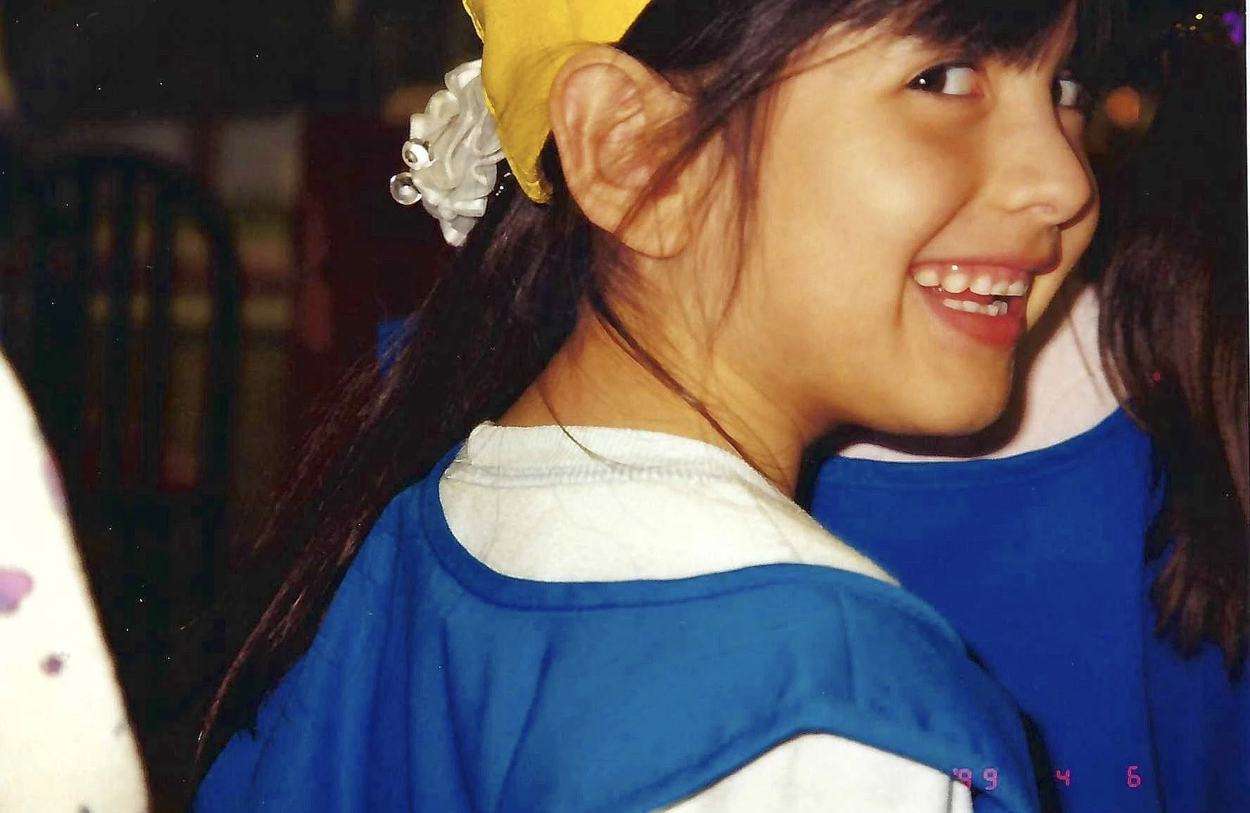 A young woman smiling with braces, wearing a yellow headband, white shirt, and blue sweater, in a social setting with blurred background.
