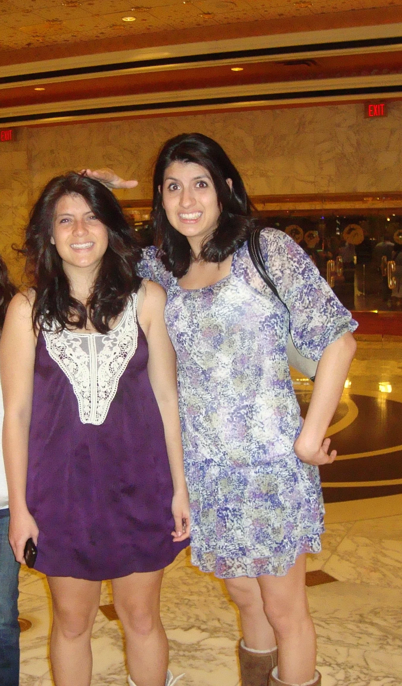 Two women standing indoors in front of a marble wall, smiling with their arms around each other. One is wearing a purple dress with lace details, and the other is wearing a floral dress with boots.