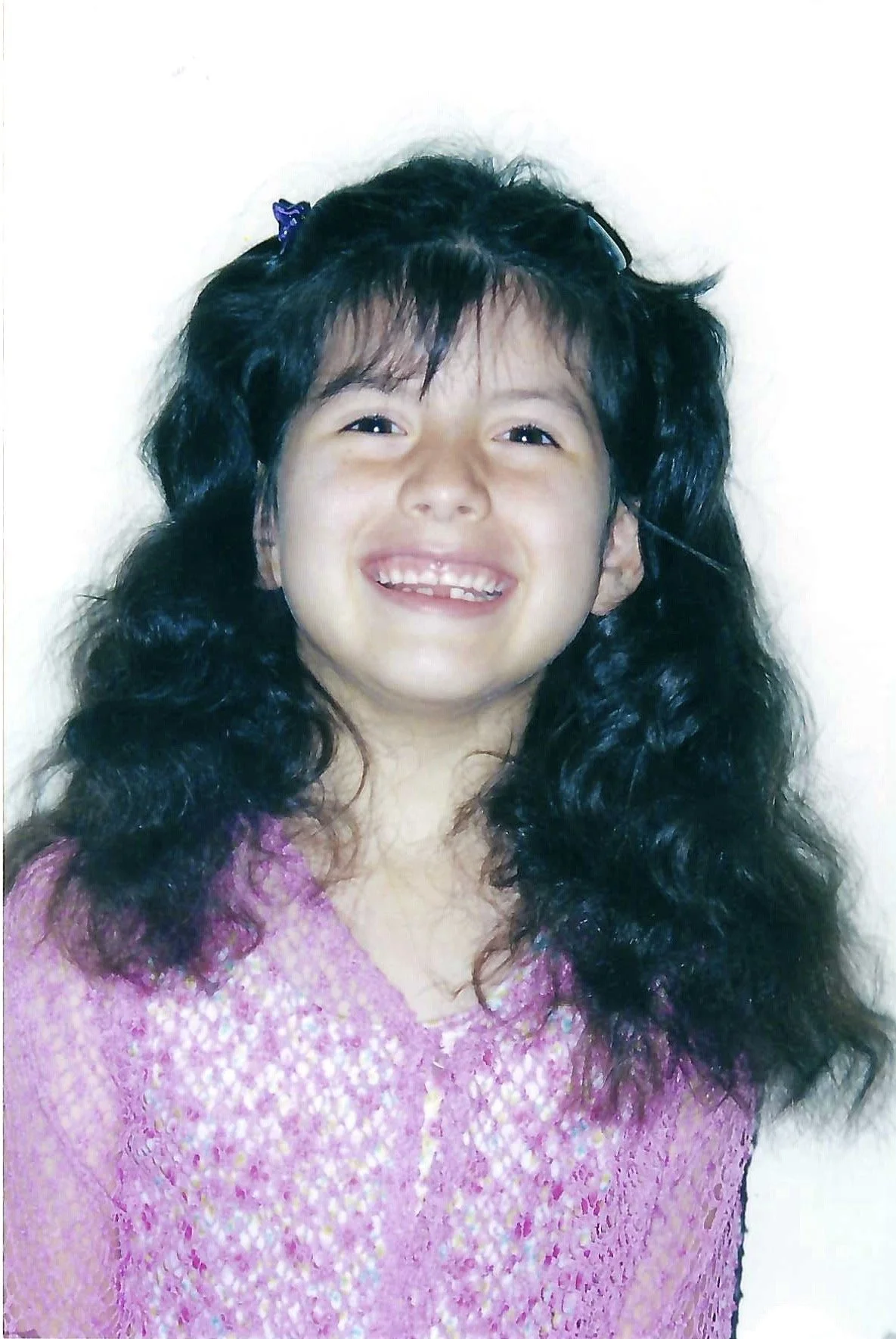 A young girl with long, curly black hair smiling widely, showing missing front teeth, wearing a pink, sparkly top, standing against a plain white background.