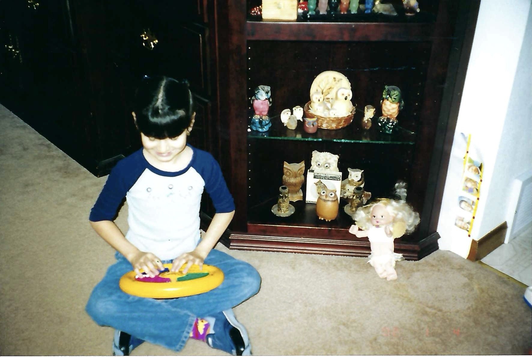 A young boy with dark hair and a bowl cut, wearing a white and blue shirt, standing and playing with a toy on a blue blanket. Behind him is a display cabinet with various ceramic animal figurines, including owls, owls, and a large bird with a second 