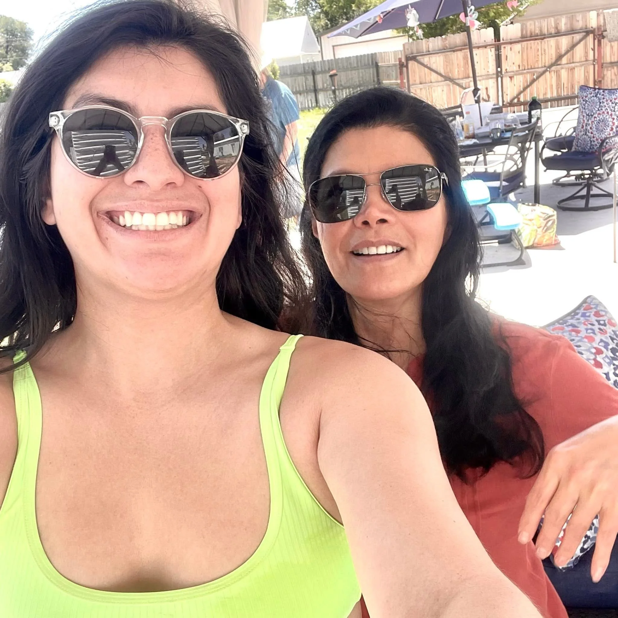 Two women smiling outdoors, both wearing sunglasses, with a wooden fence, patio table, and chairs in the background.