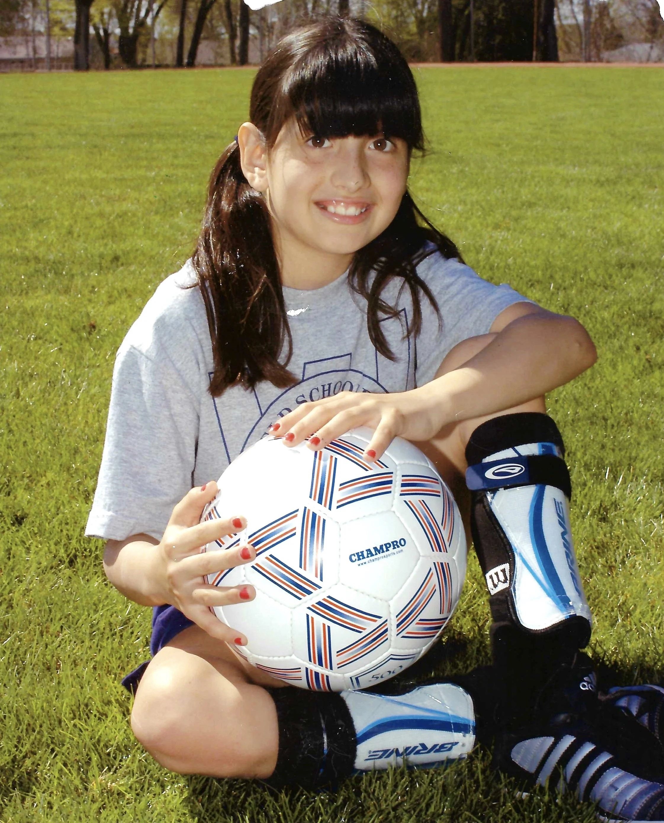A girl with dark hair and bangs sitting on grass outdoors, holding a white soccer ball with blue and orange stripes. She is smiling and wearing a gray shirt, black shorts, and soccer cleats, with a sports bottle attached to her shorts.