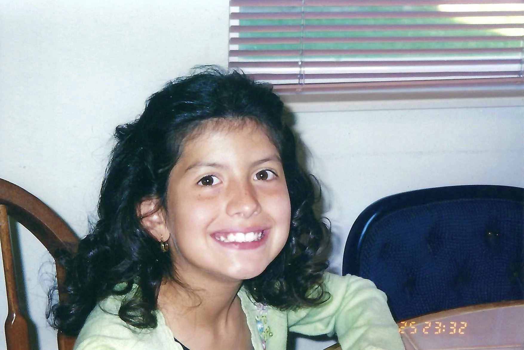 A young girl with curly black hair and a bright smile, wearing a light green top, sitting at a table with a wooden chair and a dark blue chair behind her.
