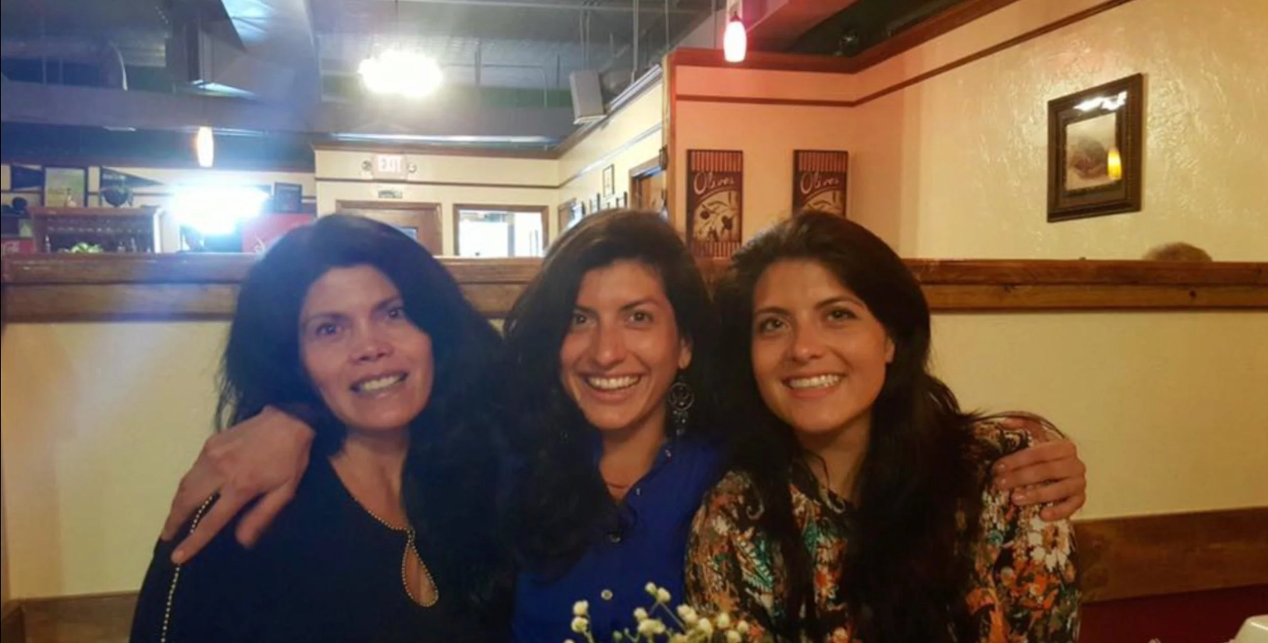 Three women smiling and sitting together in a restaurant, with arm around each other's shoulders.