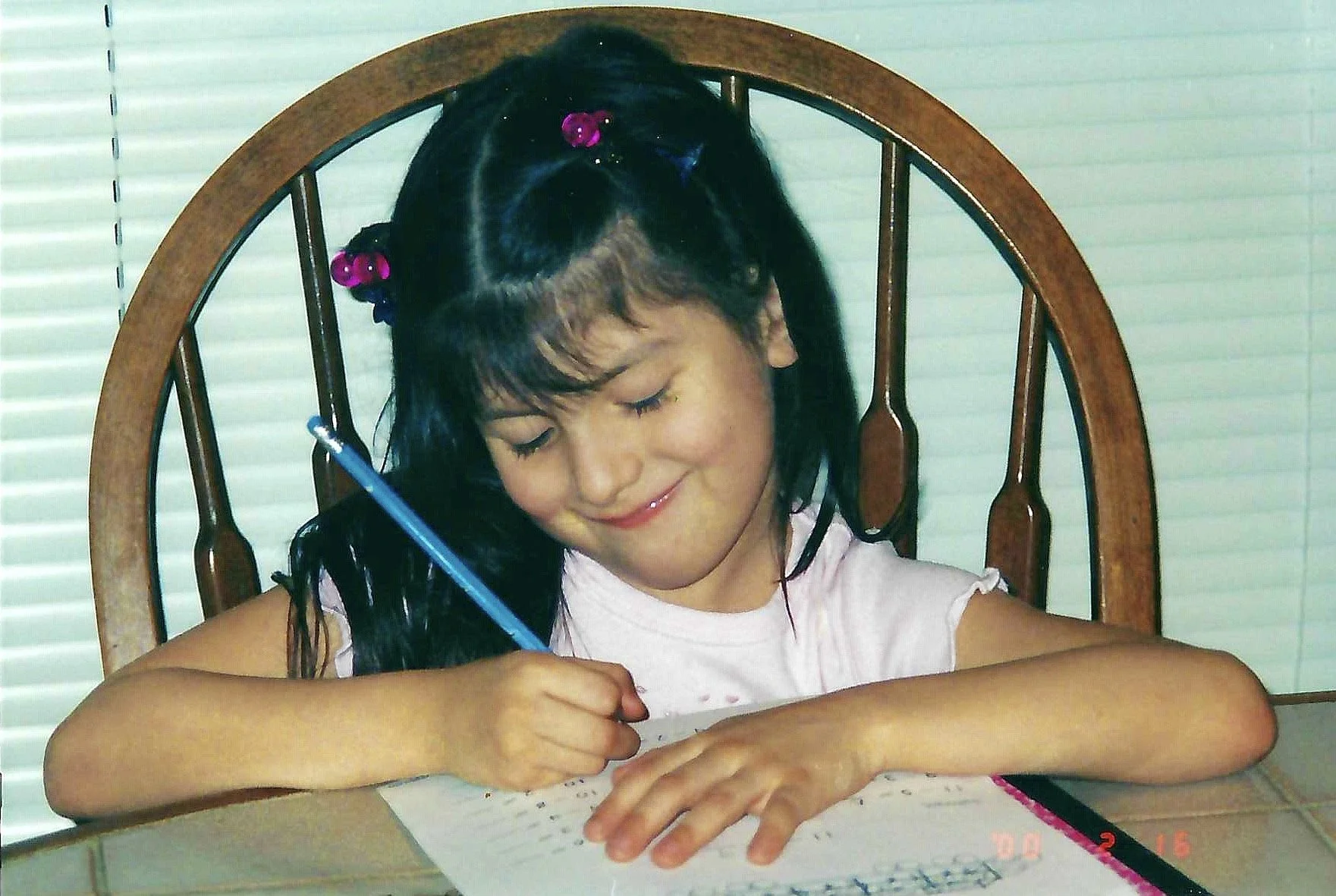 A young girl with Black hair, decorated with colorful hair clips, smiling with her eyes closed, sitting at a wooden table, holding a blue pen, writing on a sheet of paper, in front of a window with blinds.