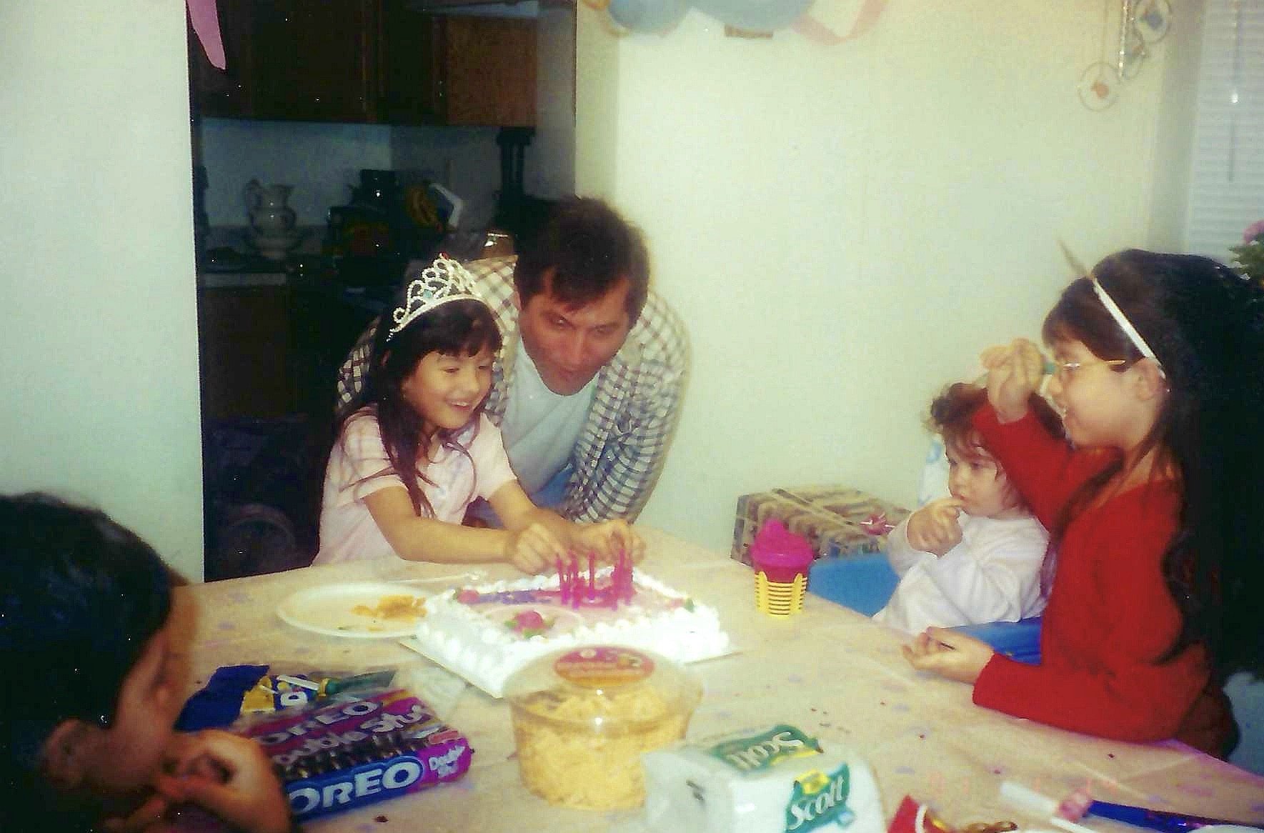 A birthday celebration with a young girl wearing a tiara, sitting at a table with a birthday cake, surrounded by family and friends. The girl is smiling, and others are gathered around, some wearing glasses, in a decorated room.