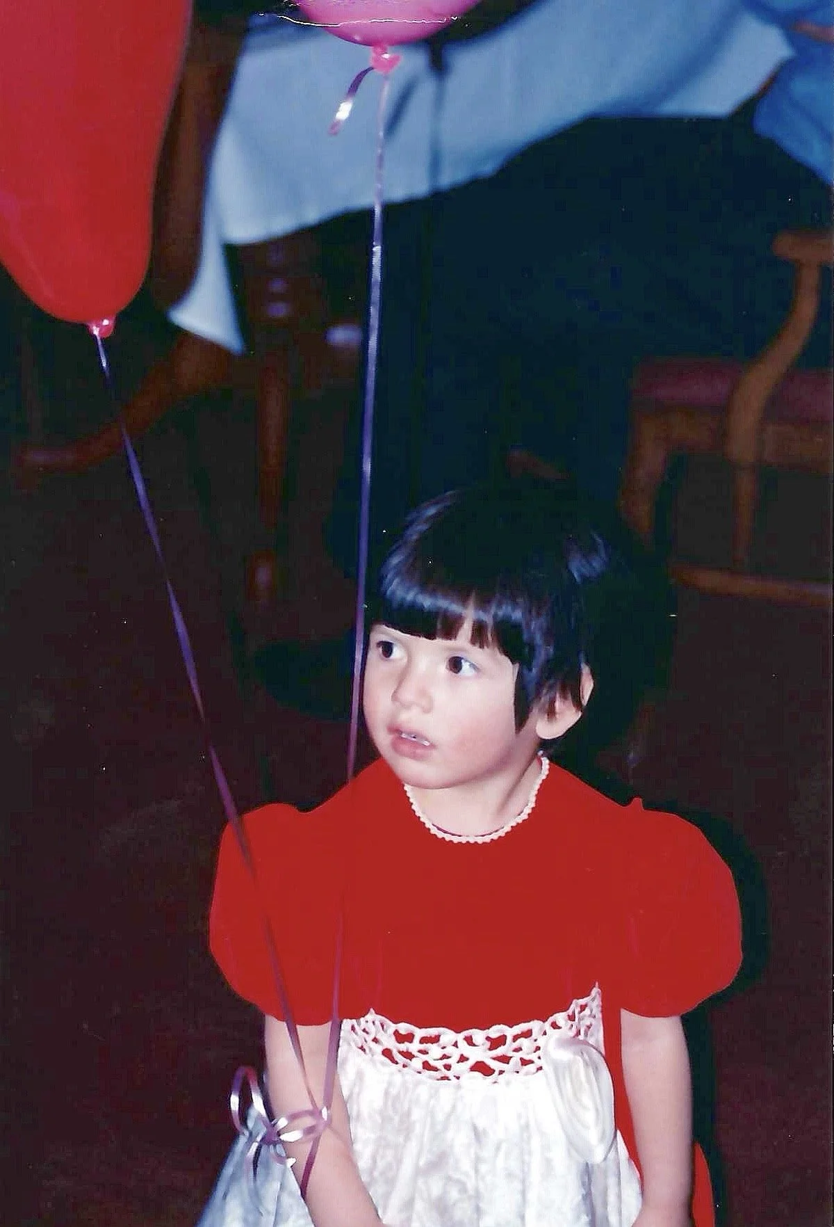 A young girl with short black hair and bangs, wearing a red top and a cream-colored skirt, is standing indoors holding purple balloons. She has a slightly surprised or curious expression on her face.