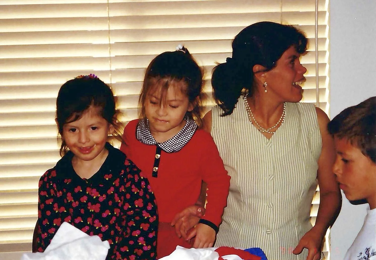 A woman with dark hair in an updo, wearing a sleeveless striped dress and jewelry, is facing right and smiling, while three young girls stand to her left, one wearing a black dress with colorful flower patterns, another in a red outfit, and the third