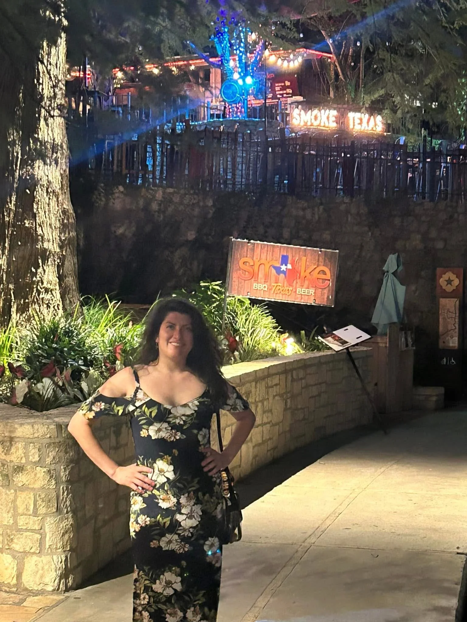 A woman with dark curly hair wearing a black floral dress standing outdoors at night, smiling with her hands on her hips in front of a brick wall, with signs for Smoke Texas BBQ, with illuminated trees and string lights in the background.