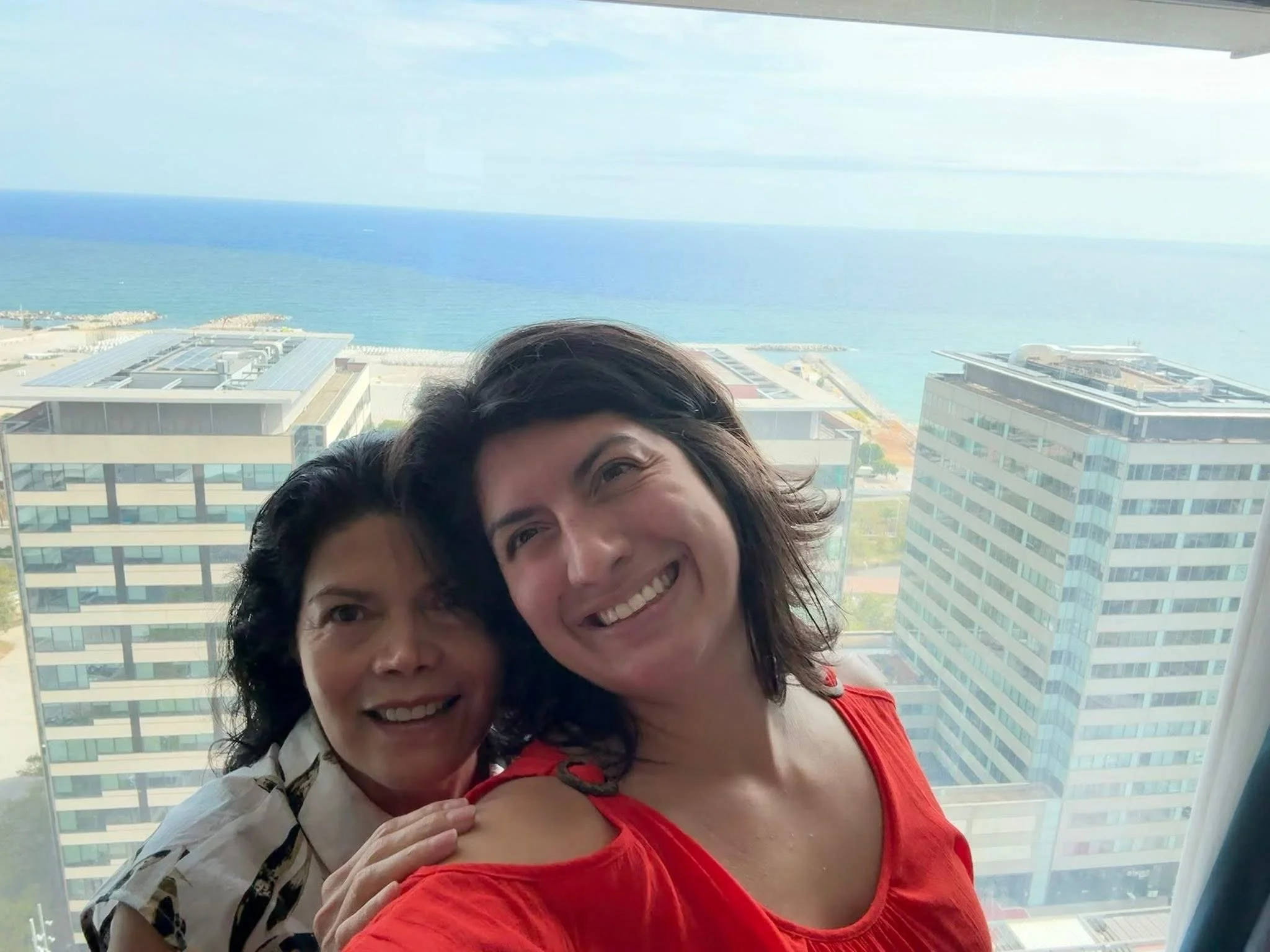 Two women smiling and hugging in front of a large window with a view of high-rise buildings and the ocean in the background.