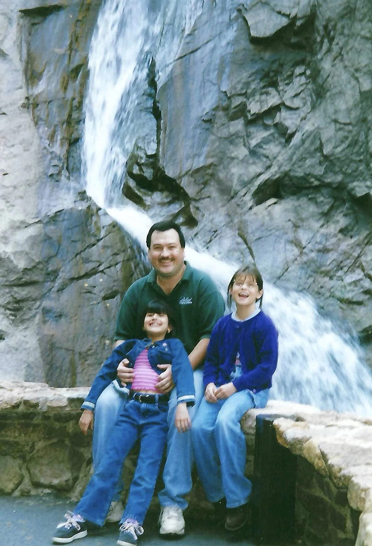 A man and two young girls sitting on a ledge in front of a waterfall.
