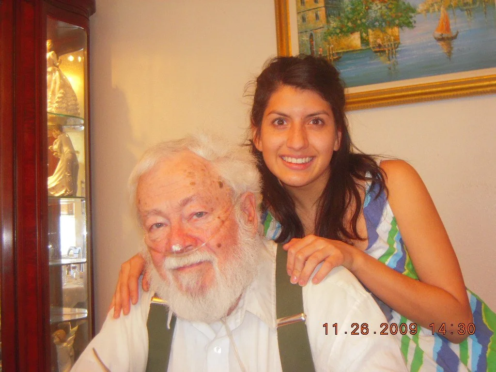 A young woman with long dark hair smiling and leaning on an elderly man with a white beard and mustache, sitting indoors with a painting of a water scene on the wall behind them.