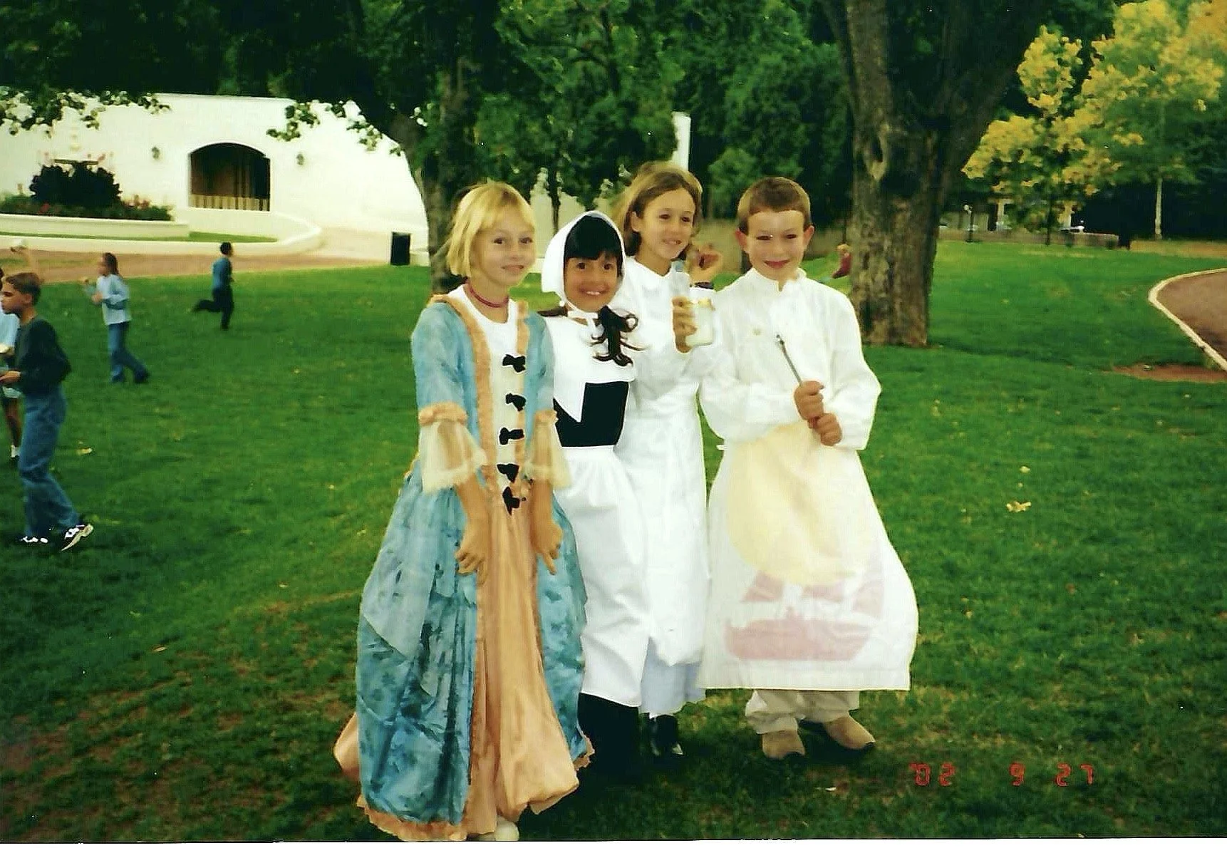 Four children dressed in costumes standing on a grassy area in a park with trees, a white building, and other children playing in the background.