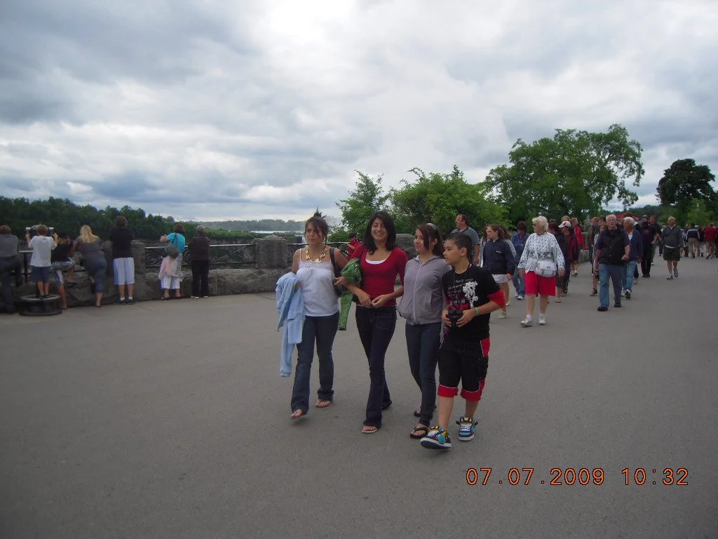 Group of people walking and standing outdoors on a cloudy day, with some looking at the view and others walking or talking, near water and trees in the background.