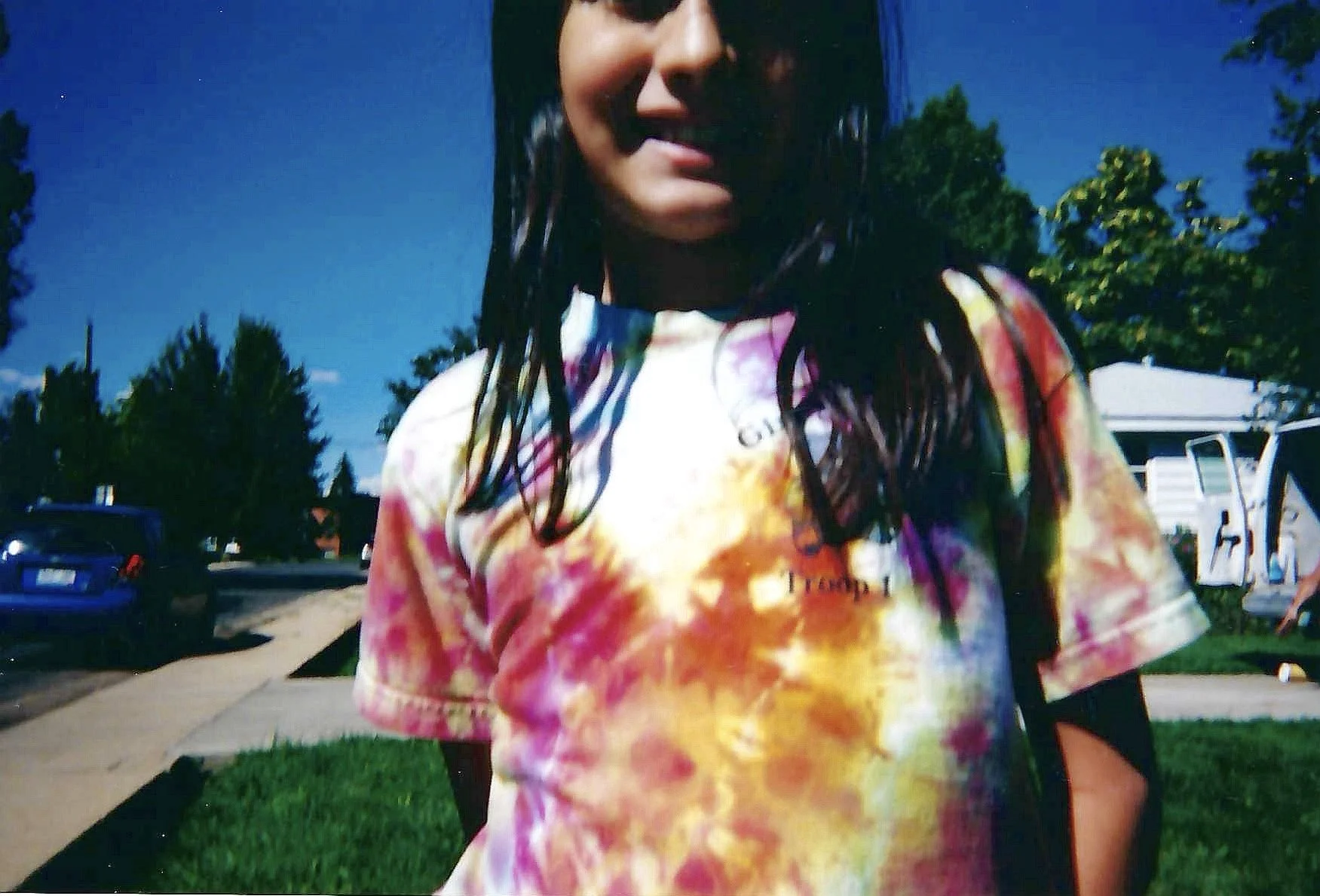 A young woman with long dark hair wearing a colorful tie-dye t-shirt, standing outside on a sunny day with green trees and a clear blue sky in the background.