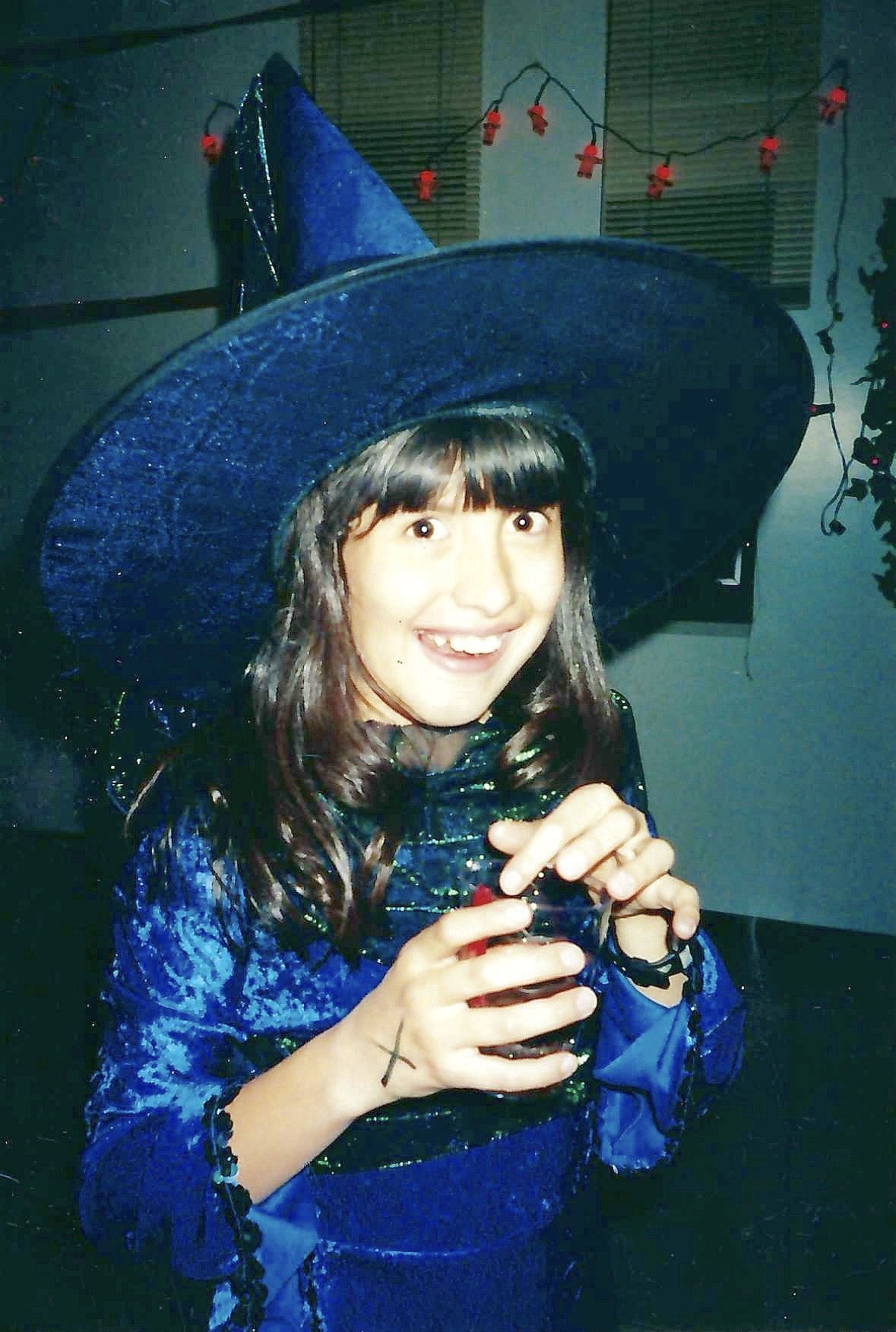 A young woman wearing a large, sparkly blue witch hat and matching shiny blue costume, smiling and holding a drink in a festive indoor setting with holiday lights in the background.