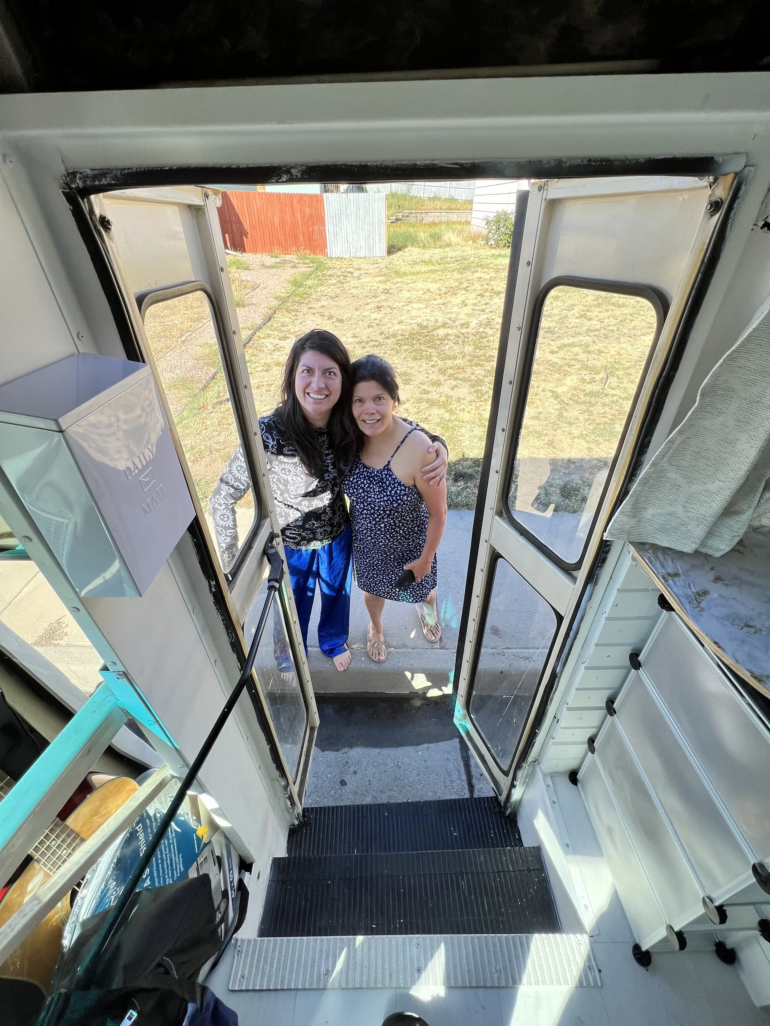 Two women standing outside an open food truck door, smiling and hugging each other, with a grassy yard and red and blue fencing in the background.