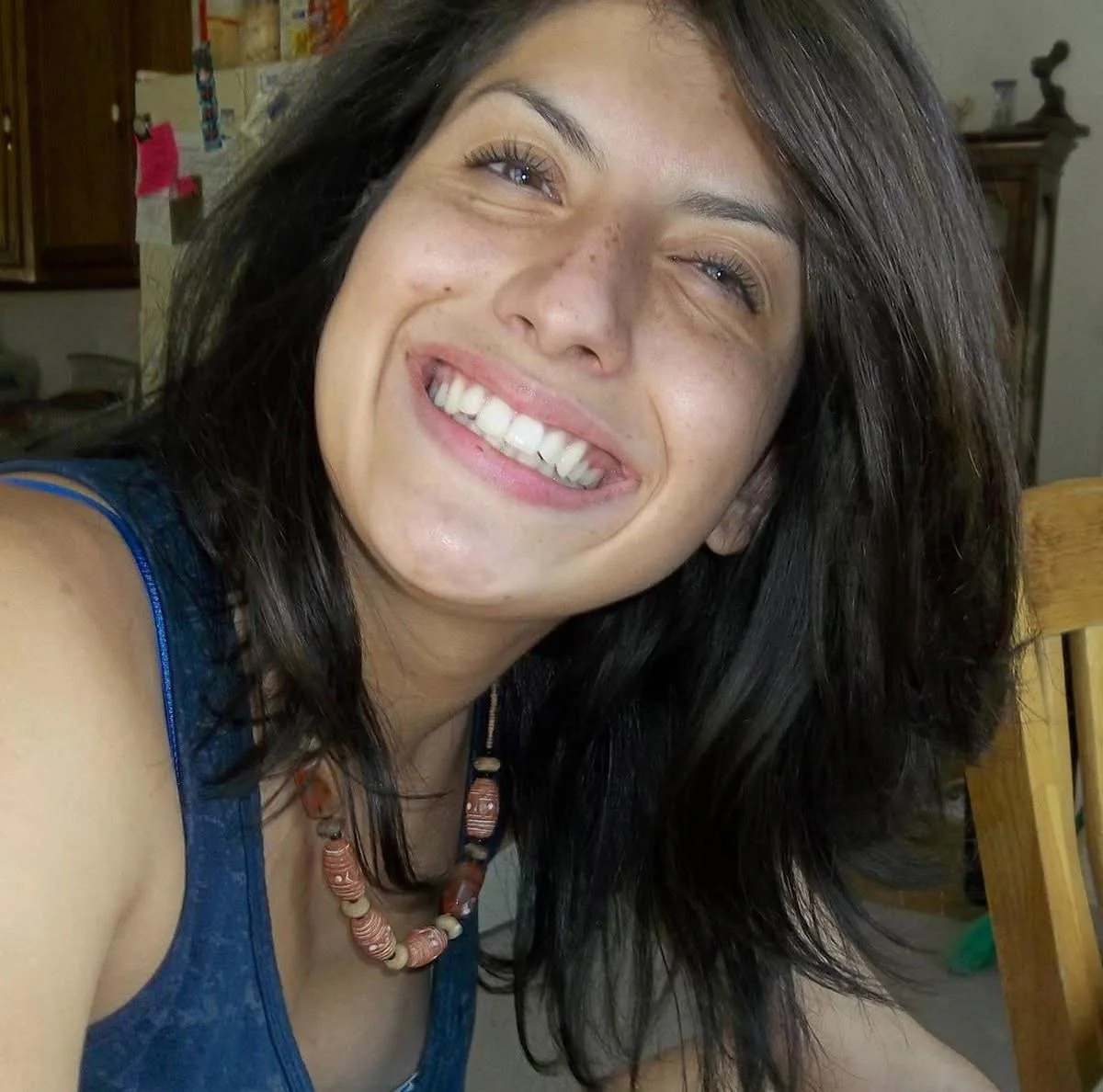 A young woman with dark hair, smiling widely, showing her teeth, in an indoor setting with wooden furniture and cabinets in the background.