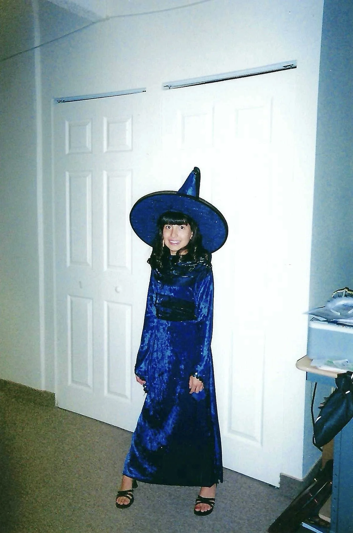 A young girl dressed as a witch, wearing a blue velvet gown and a matching pointed hat, standing in front of white double doors in a room.