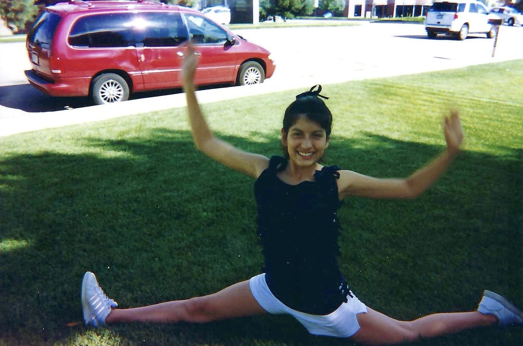 A young girl is doing a front split on the grass, smiling with arms raised. She is wearing a black top, white shorts, and white sneakers. There are cars parked on a street in the background under a sunny sky.