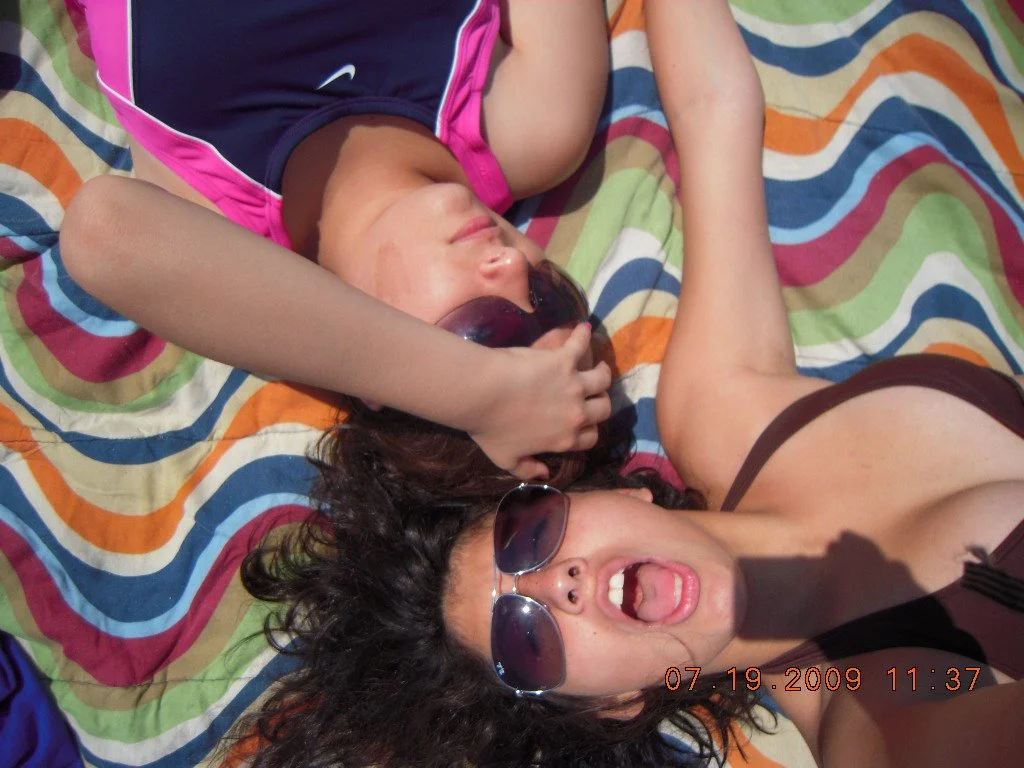 Two women lying on a colorful striped blanket, wearing sunglasses, with one woman sticking her tongue out and the other resting her arm over her face.