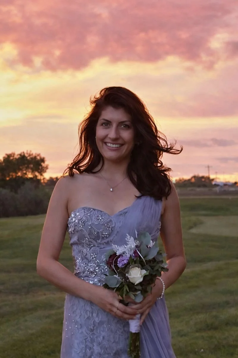 Kathy Endicott is standing holding a flower arrangement. This picture was taken at her sister's wedding. She is wearing a greyish/purple dress and looks beautiful.