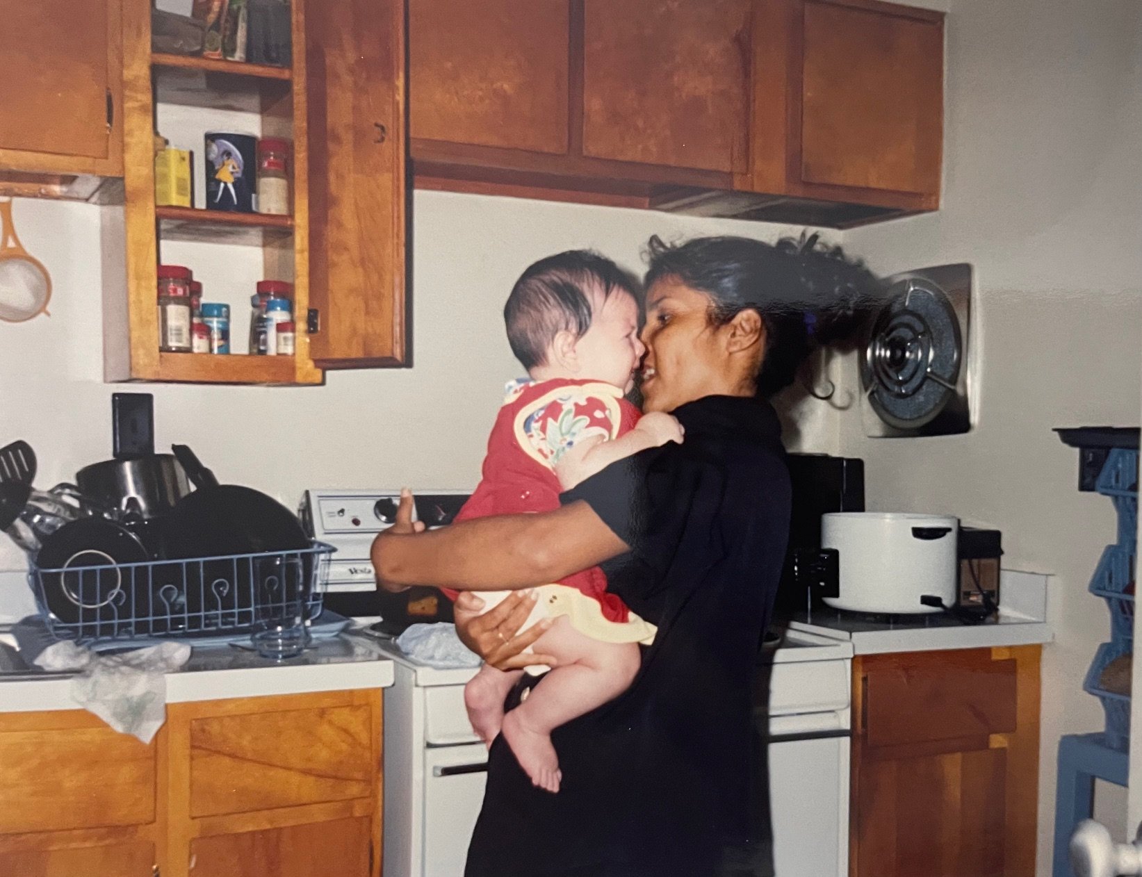 A woman holding a young child in her arms in a kitchen, smiling as they touch noses. The kitchen has wooden cabinets, a stove, and various kitchen items on the counter and shelves.