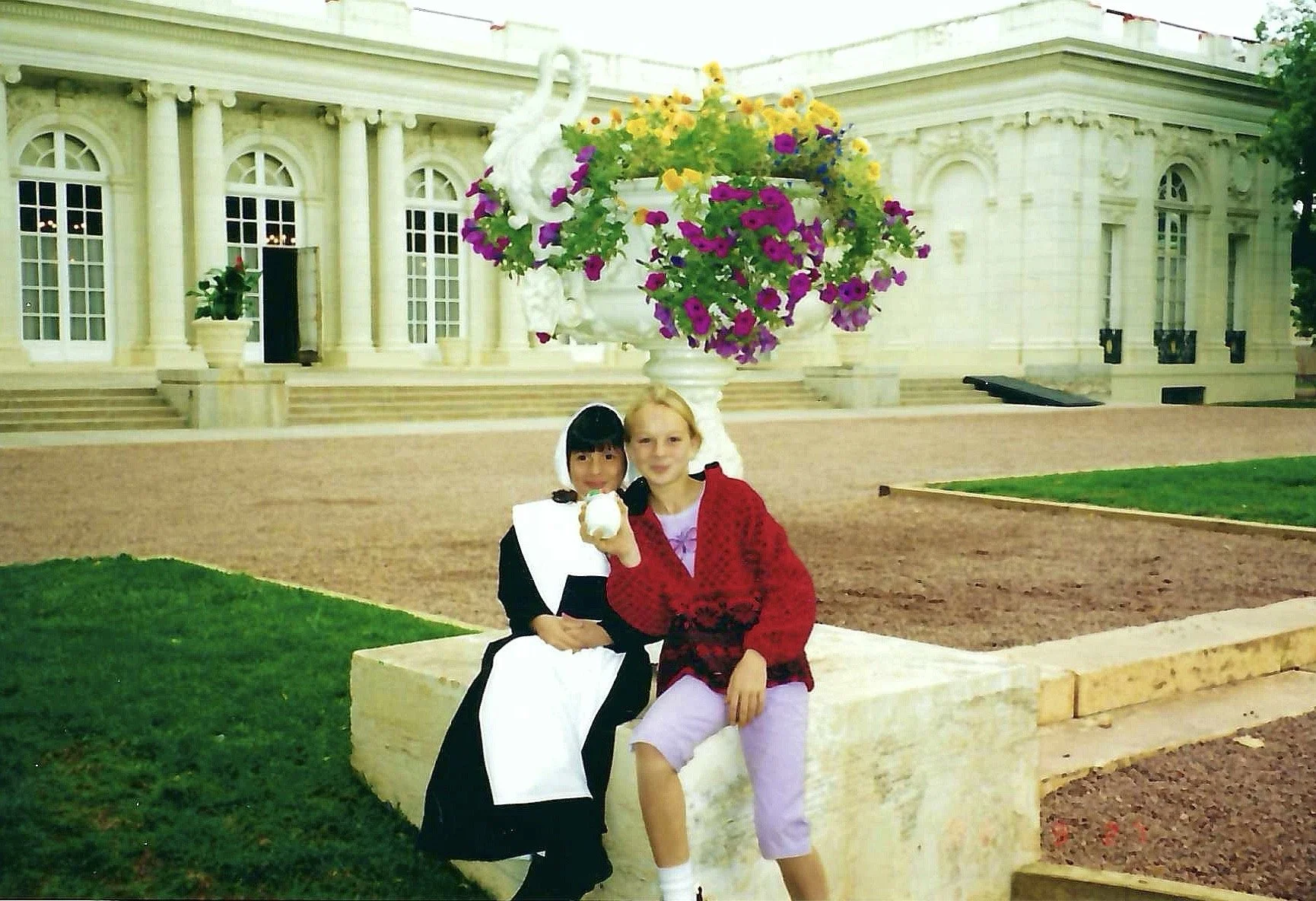 Two young girls sitting on a stone bench in front of a large white building with columns, steps, and arched windows, adorned with a decorative floral arrangement and a white swan sculpture.