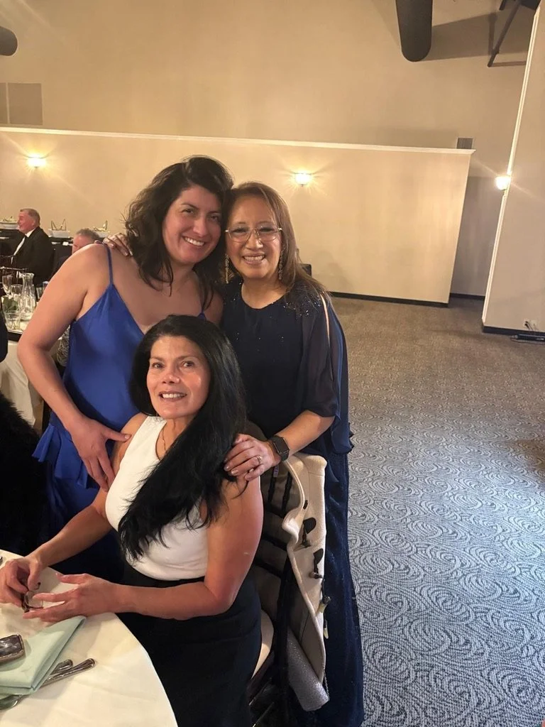 Three women at a formal event, two are standing behind, one is seated, all smiling at the camera. The woman standing on the left wears a blue dress, the woman standing on the right wears a dark blue dress, and the woman seated wears a white top and d