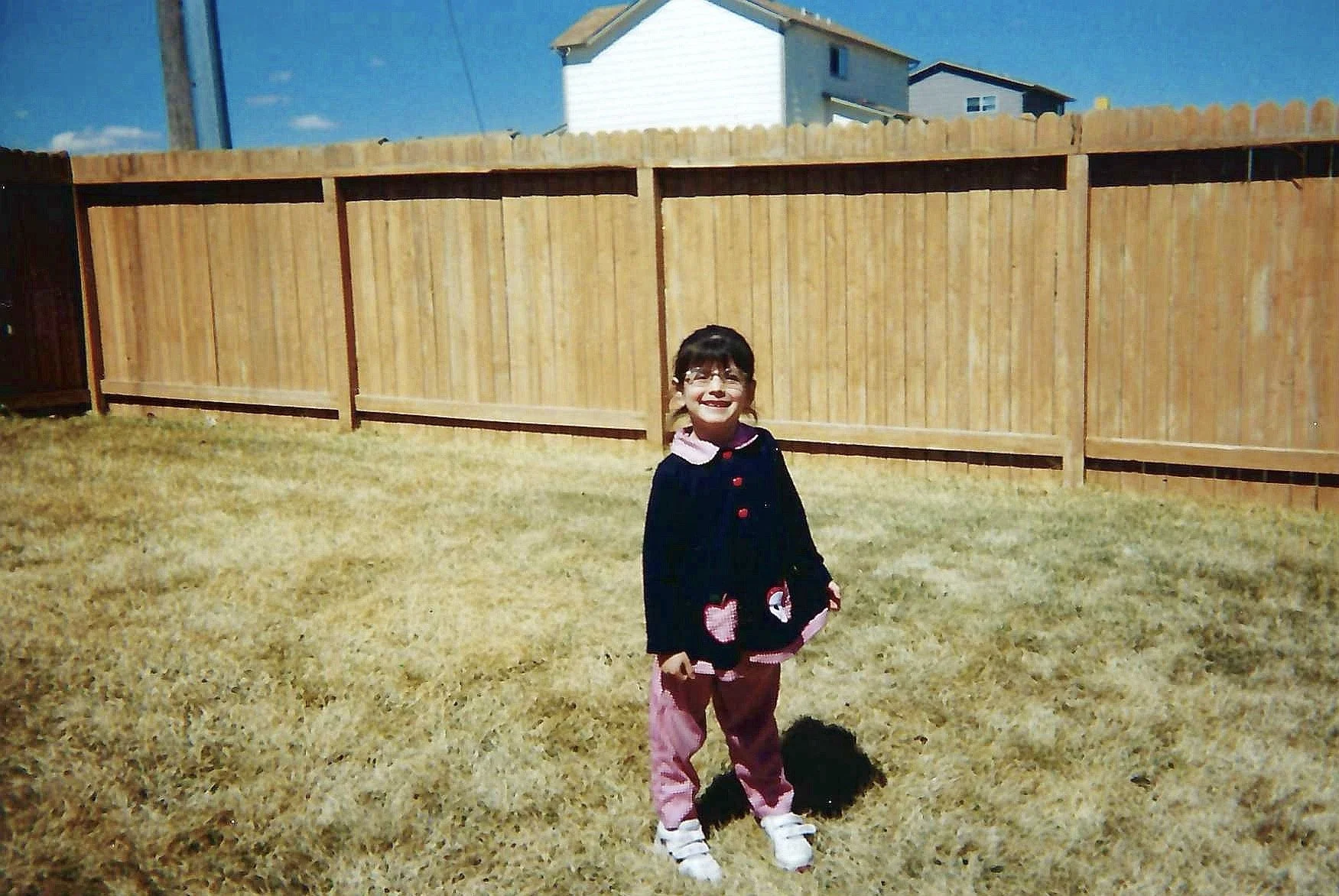 A young girl with glasses and a black jacket standing on a yellowed grass lawn in front of a wooden fence, with houses and a blue sky in the background.