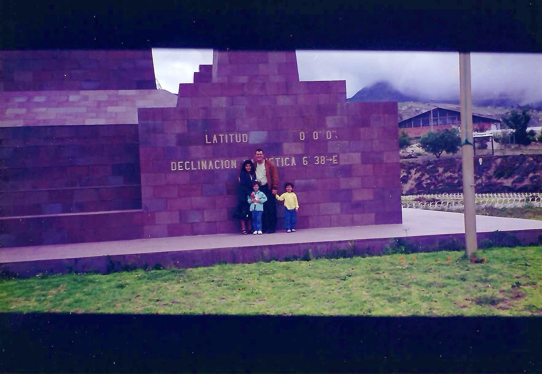 Group of four people, including two children, standing in front of a brick wall with altitude and latitude coordinates, with mountains, trees, and a building in the background under a cloudy sky.