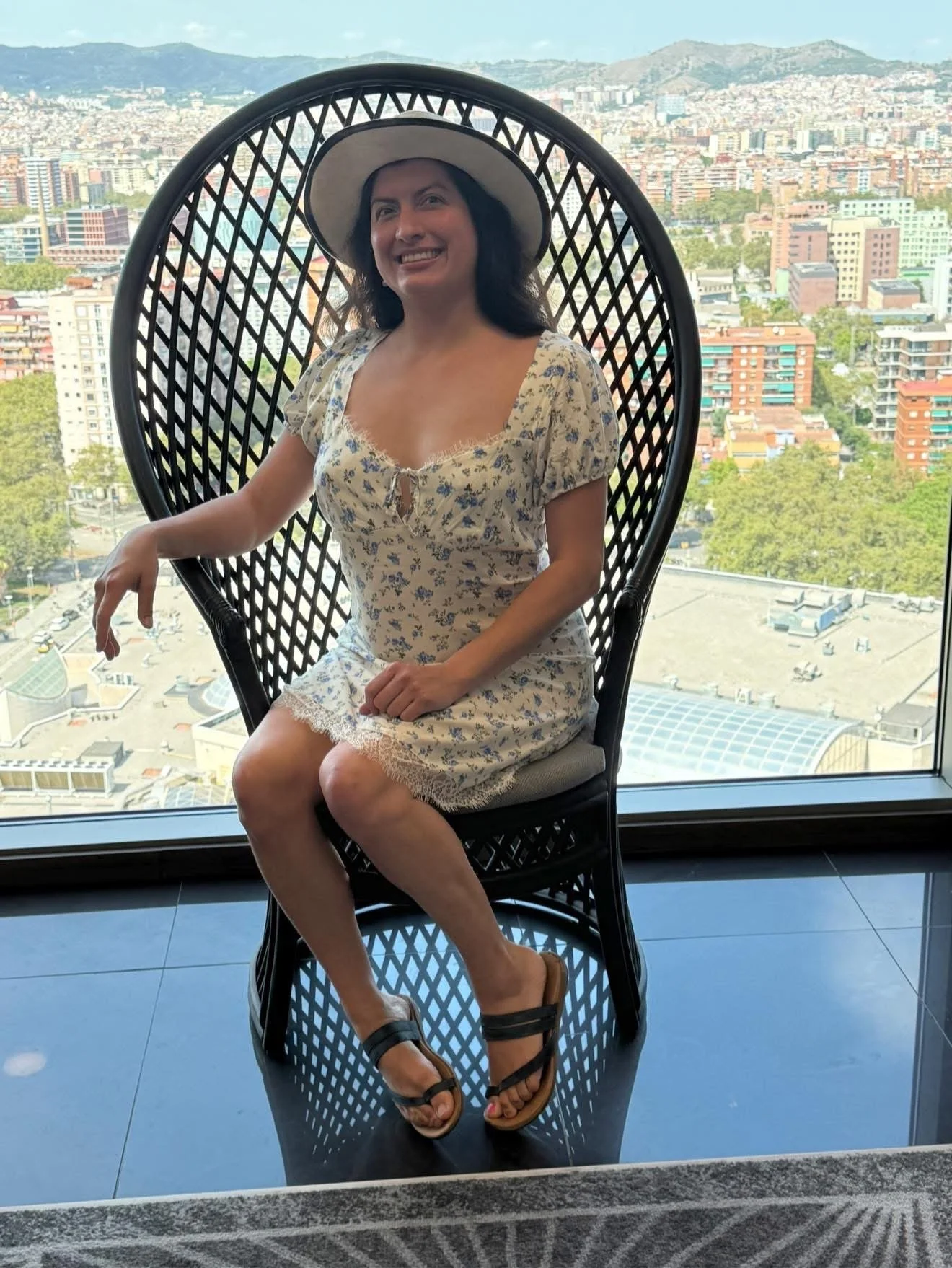 A woman sitting on a decorative black wicker chair with a large circular backrest, positioned indoors near a large window overlooking a cityscape with tall buildings and mountains in the background. She is smiling, wearing a light floral dress, a wid