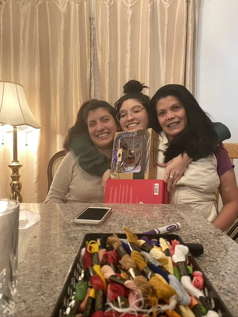 Three women sitting at a table, smiling and hugging, with a gift bag on the table. In the foreground, there is a box of embroidery floss and a smartphone.