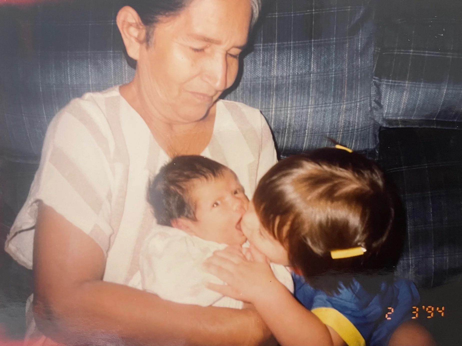 A woman sitting on a couch holding a young child, with another person in front of them. The woman has dark hair pulled back and is wearing a striped shirt. The small child has dark hair and is looking at the person in front of them. The person in fro