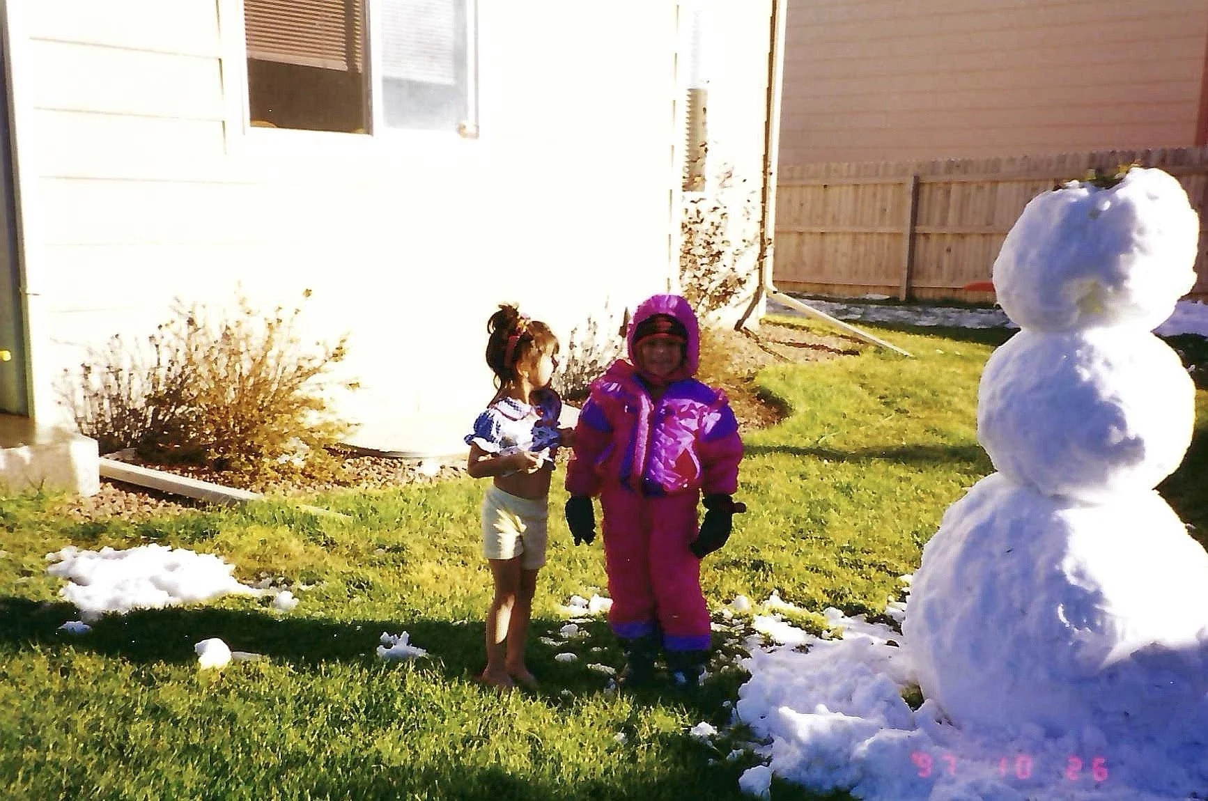 Two children stand outside on a sunny day near a three-ball snowman on the grass. One child is wearing a purple winter suit, hat, and gloves, while the other is dressed in shorts, a top, and a headband. There is snow on the ground and a house with a 