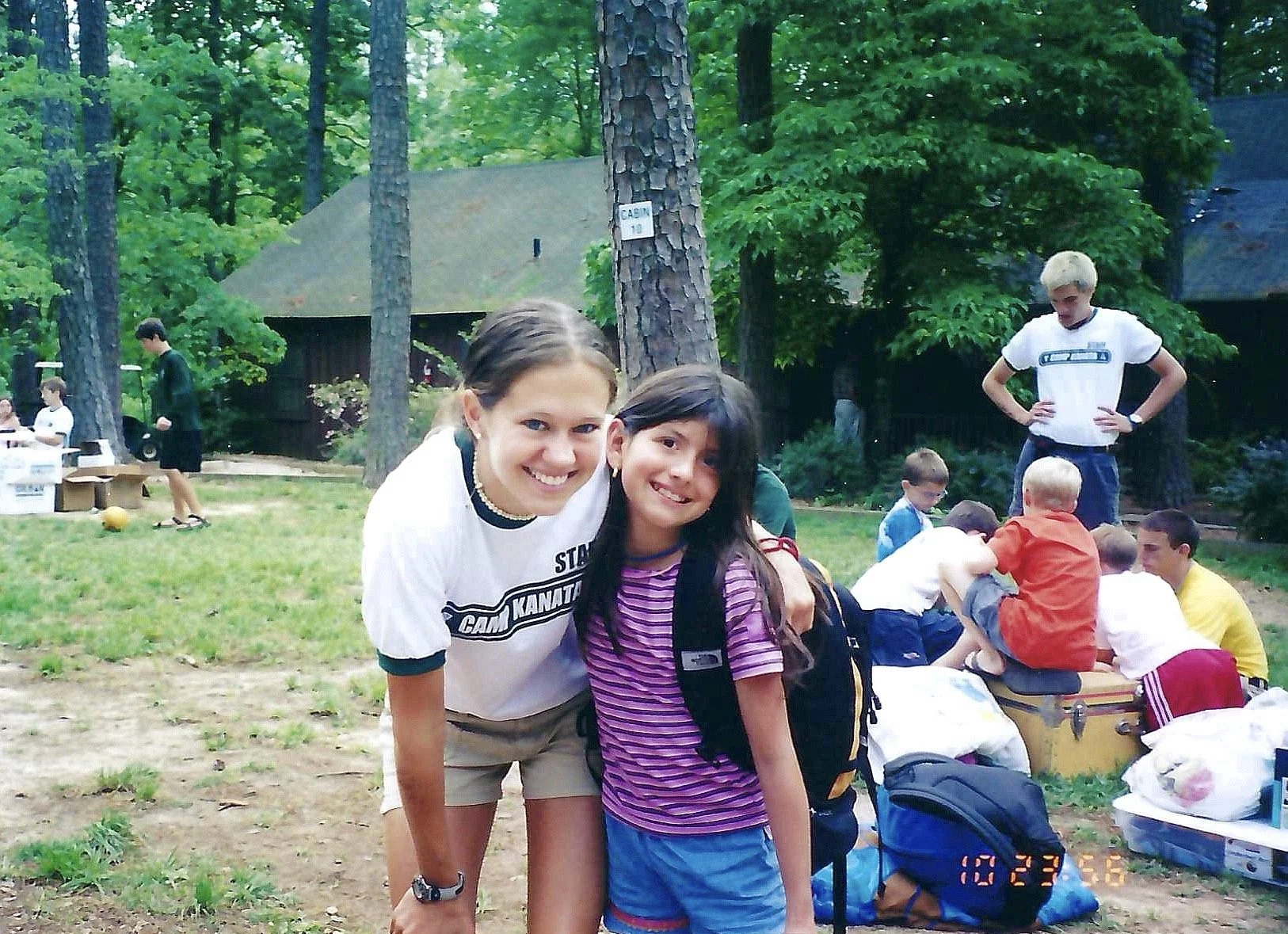 A group of children and teenagers gathered outdoors in a wooded area, with two girls smiling and posing for the camera in the foreground, and other children and teens in the background engaged in activities.