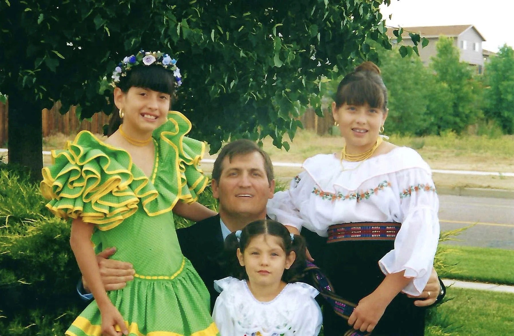A group of people, including two young girls dressed in traditional Mexican folk dresses, posing outdoors in front of a leafy tree and residential buildings.