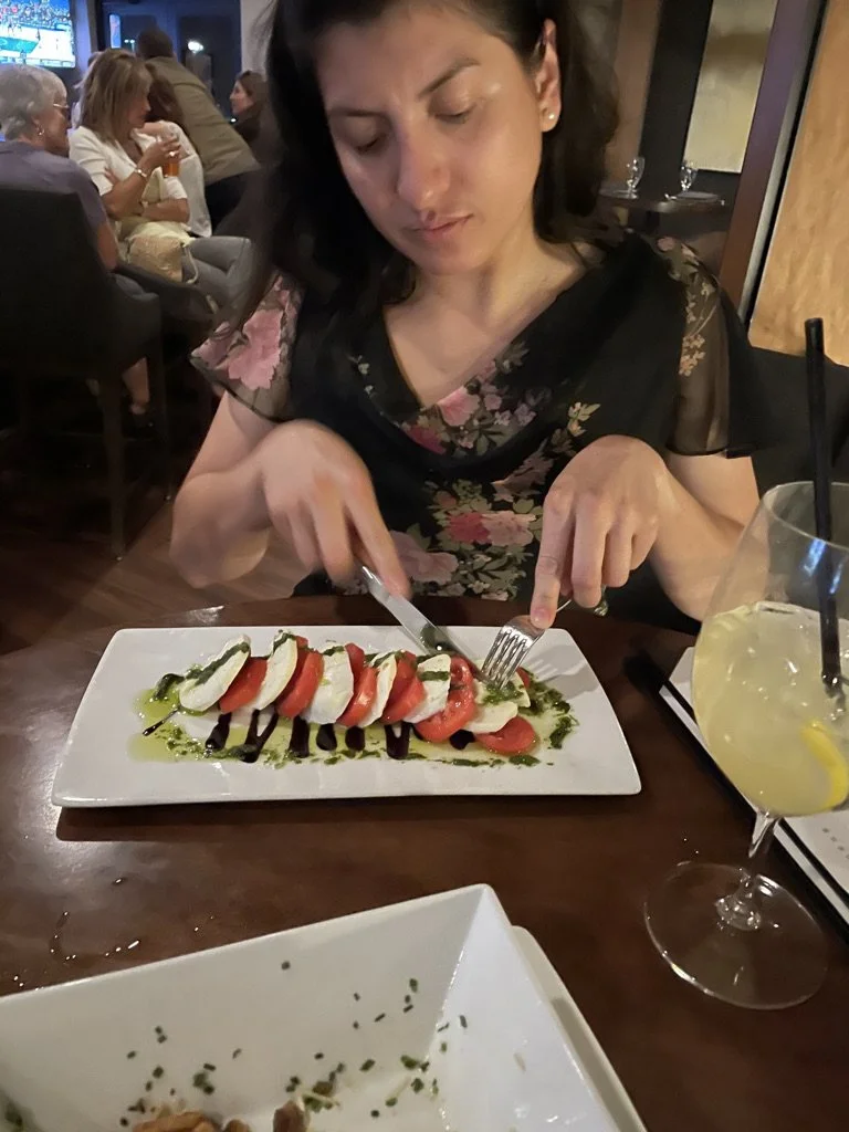 A woman in a black floral dress eating a Caprese salad with tomatoes, mozzarella, and basil, drizzled with balsamic vinegar, in a restaurant with other diners in the background. A glass of lemonade with lemon slice and straw is on the table.