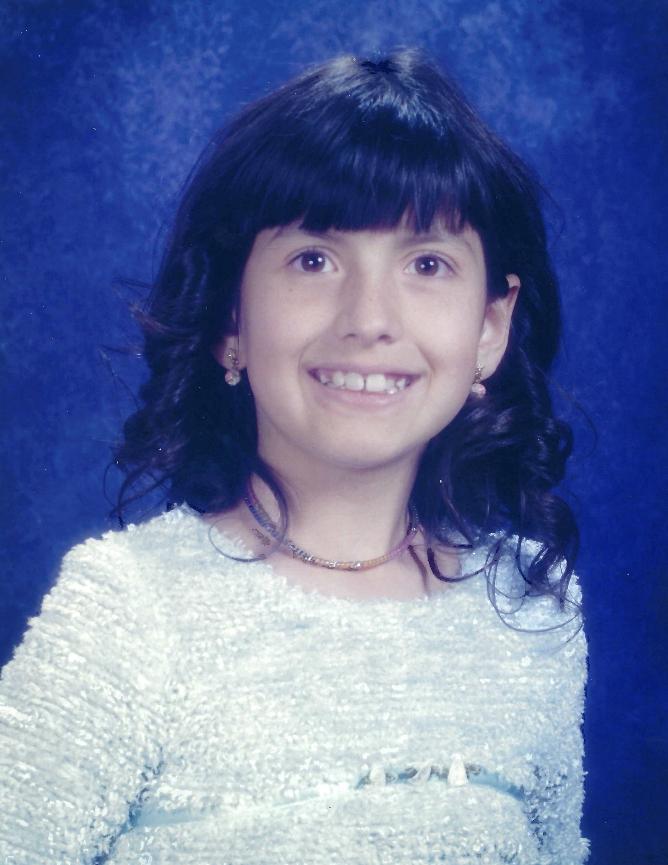 A woman with dark hair and earrings smiling, wearing a white textured sweater and jewelry, against a blue background.