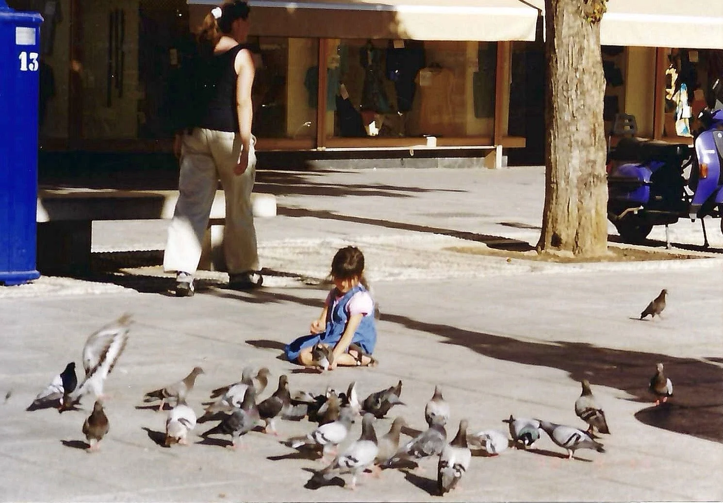 A young girl sitting on the sidewalk surrounded by pigeons, with an adult woman standing nearby, in an outdoor urban setting. There are trees, a parked scooter, and a building in the background.