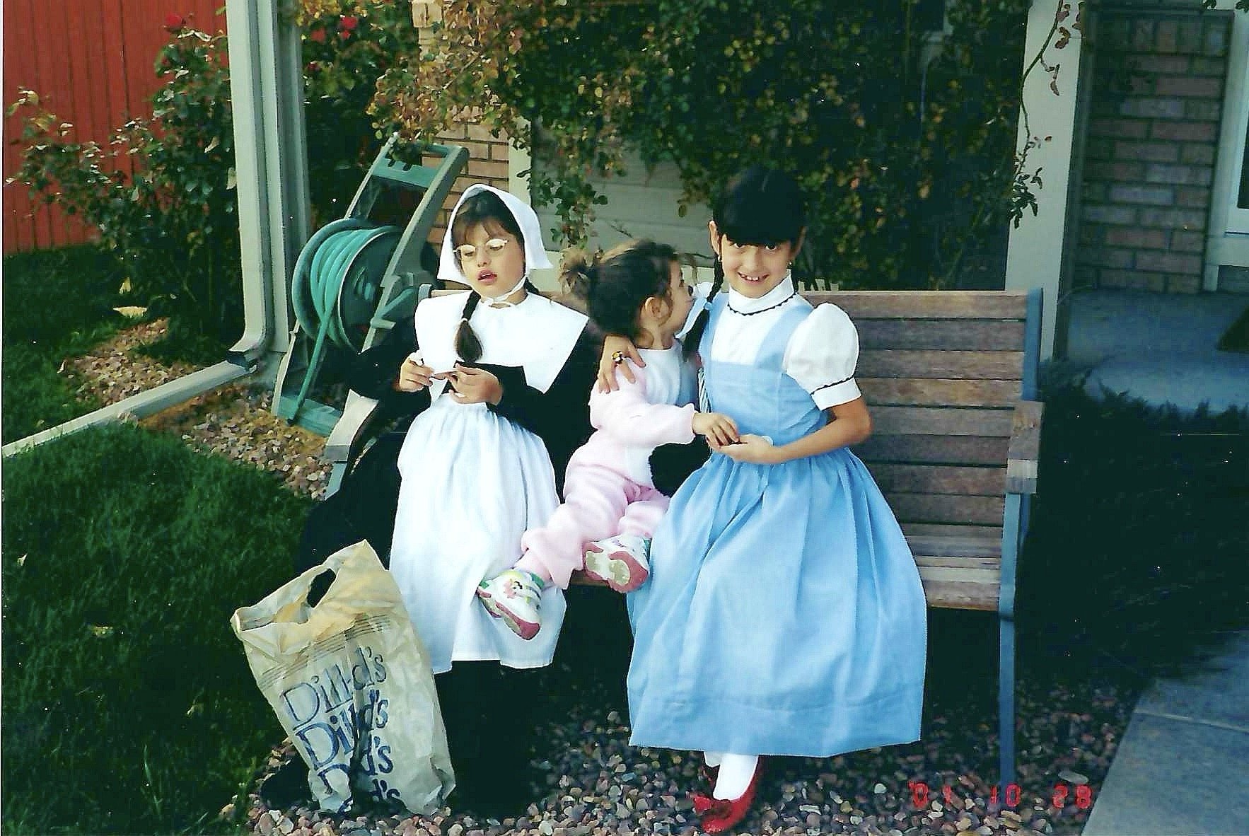 Four children dressed in costumes sitting on a wooden bench outdoors, with a garden hose reel and bushes in the background, and a paper bag on the ground.