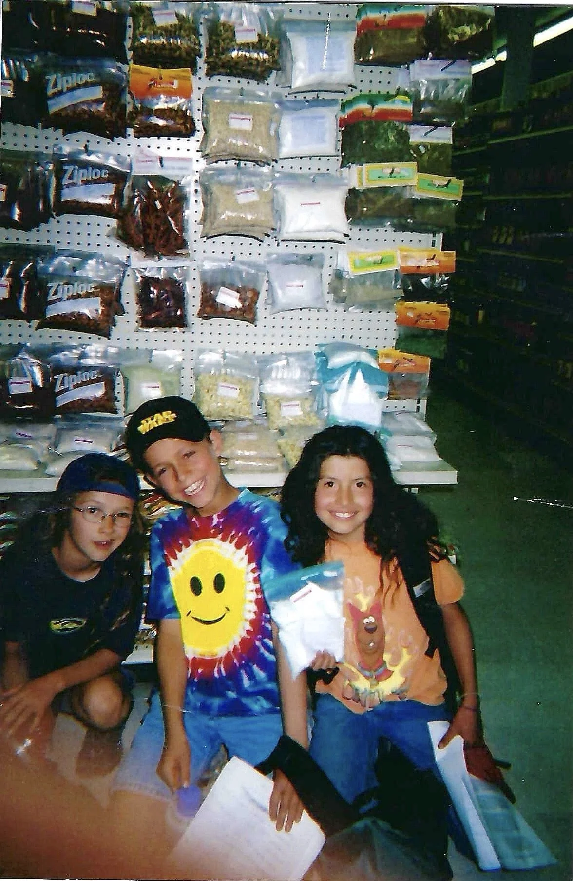 Three children standing in front of a store shelf filled with packaged food items, smiling and holding shopping items.